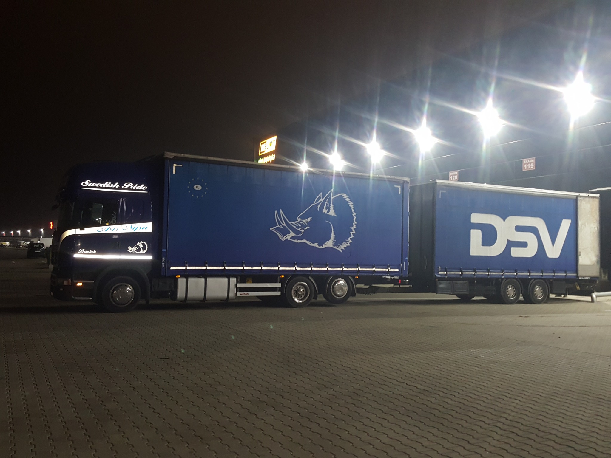 Ciężarówka z naczepą, w barwach niebieskich z logo firmowym przedstawiającym głowę zwierzęcia, zaparkowana na placu w nocy, oświetlona przez liczne lampy.