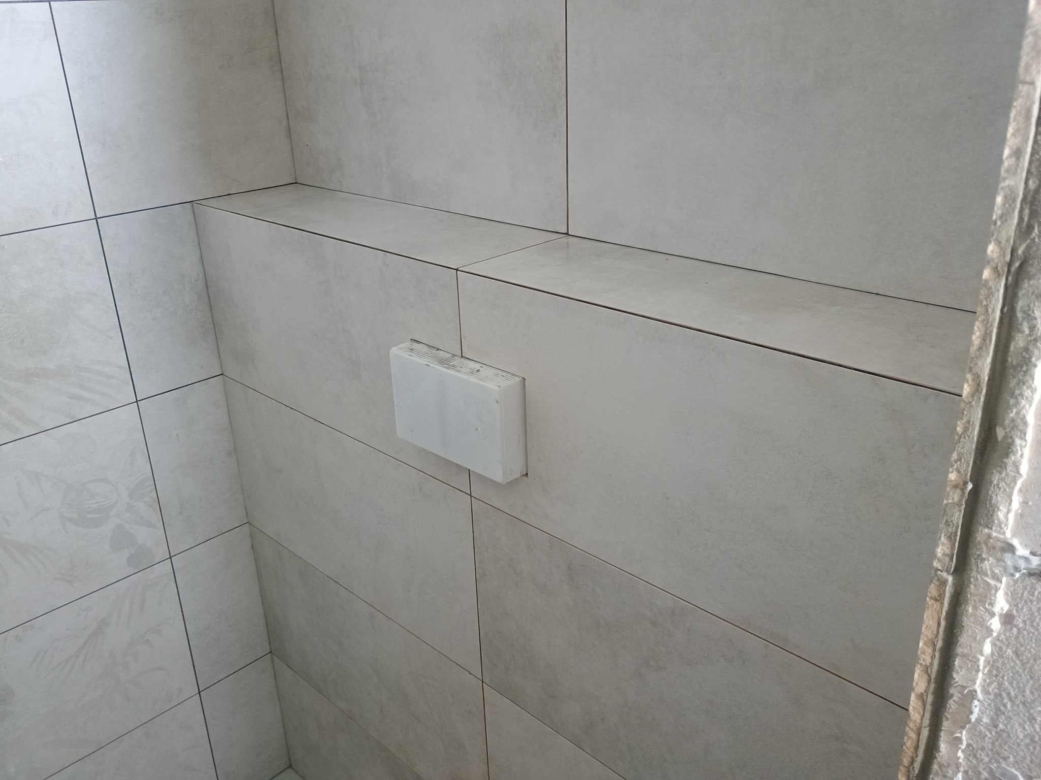 Wykończona łazienka z jasnymi, kwadratowymi płytkami ceramicznymi na ścianach i półce. Widoczny fragment ściany w surowym stanie. Minimalistyczny design.