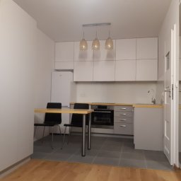 RAJ BUD - Jasna kuchnia w minimalistycznym stylu z białymi szafkami, szarymi blatami i podłogą, oświetlona trzema wiszącymi lampami z bursztynowego szkła, z widocznym stołem z dwoma krzesłami i częściowo...