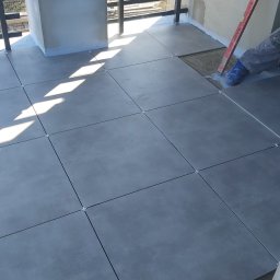 RAJ BUD - Układanie szarych płytek podłogowych na balkonie, widoczne krzyżyki dystansowe, poziomica i fragment buta glazurnika.