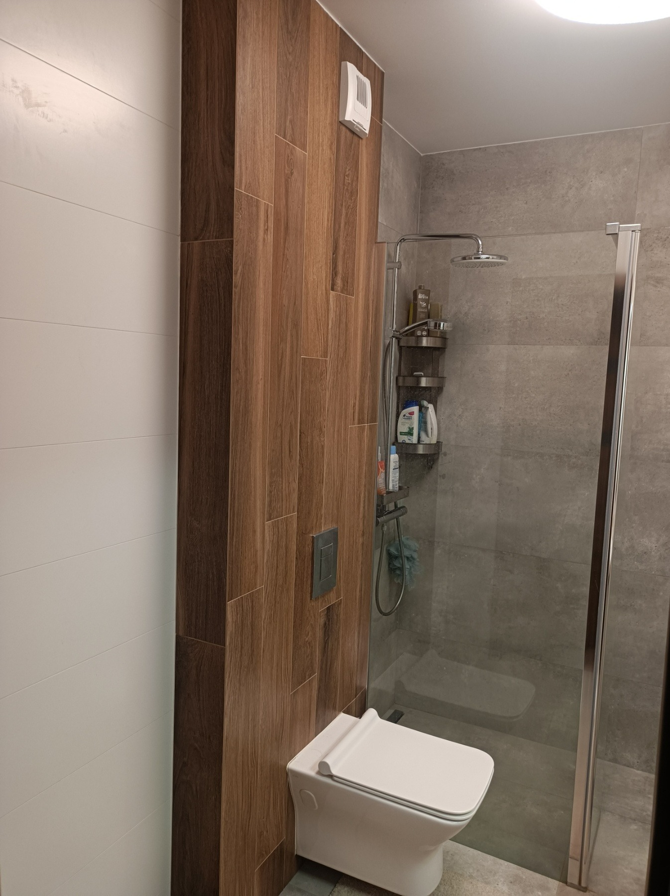 Nowoczesna łazienka z kabiną prysznicową, białą toaletą wiszącą i ścianą wyłożoną płytkami imitującymi drewno, kontrastującą z białą ścianą z poziomymi płytkami.