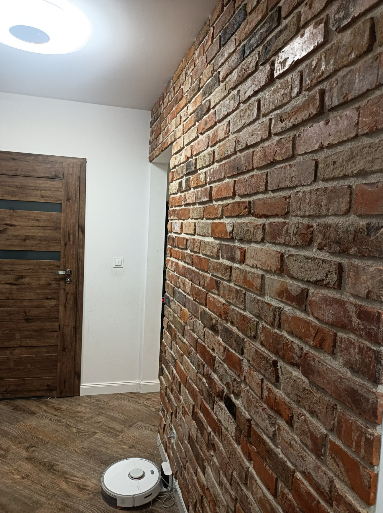 Rustykalna ściana z czerwonej cegły w mieszkaniu w Otwocku, z widoczną drewnianą podłogą i drzwiami z przeszkleniami, oraz robotem sprzątającym przy ścianie.