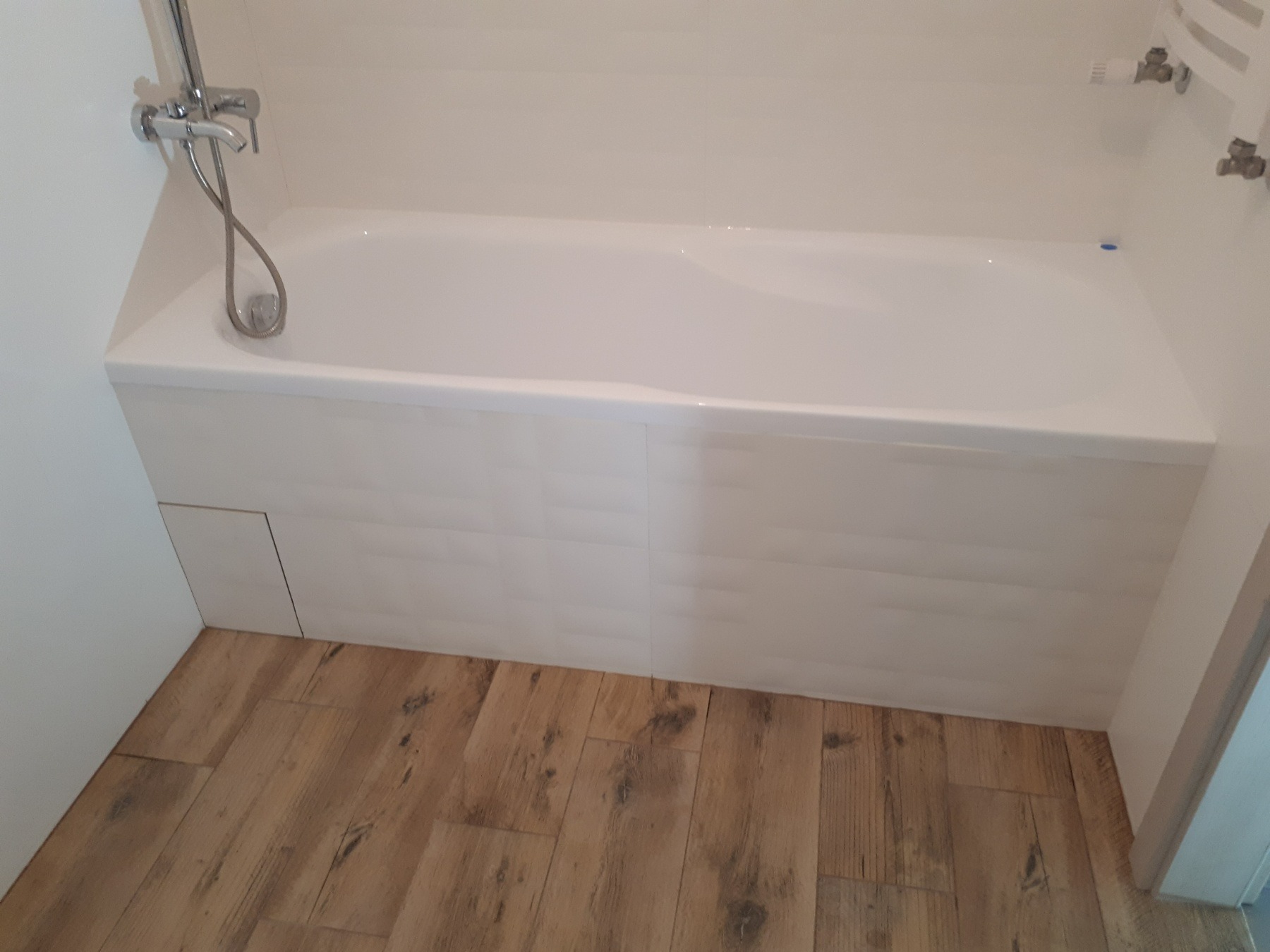 Biała wanna z baterią, obudowana płytkami imitującymi cegłę, z drewnopodobną podłogą, widok z góry.