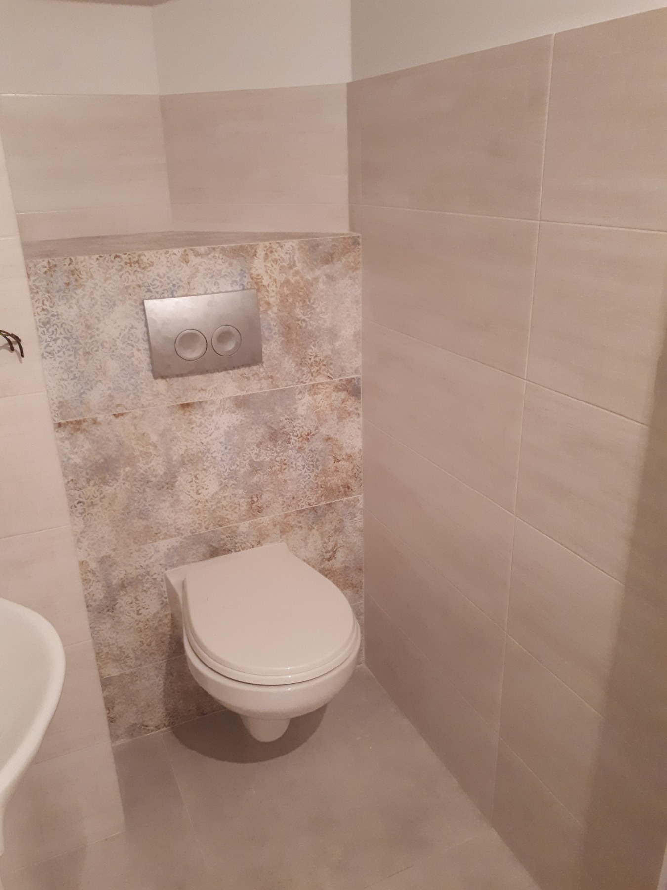 Zawieszona toaleta z chromowanym przyciskiem spłukującym na ścianie wyłożonej dekoracyjnymi płytkami w neutralnych barwach, sąsiadującej z jasnymi, gładkimi płytkami.