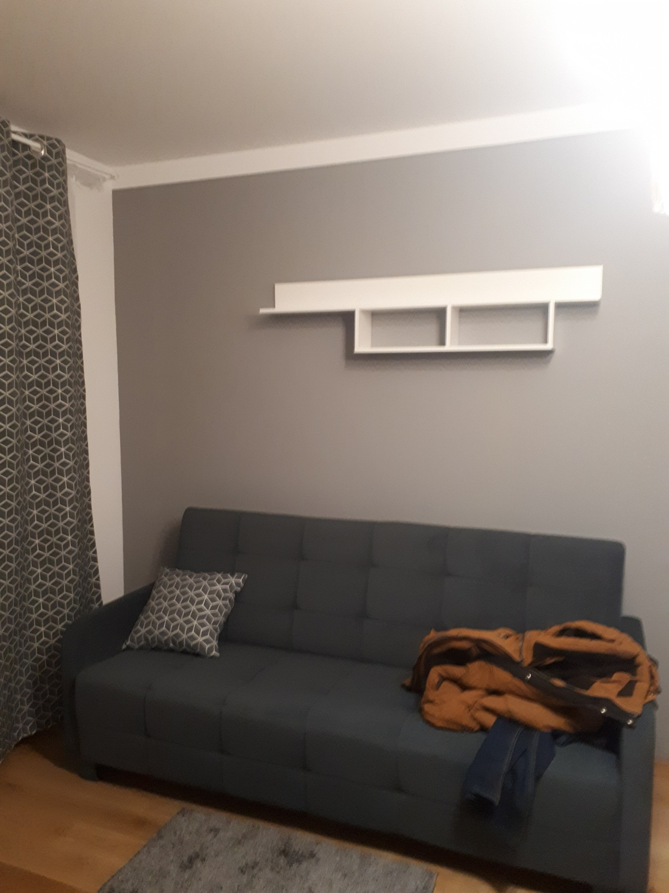 Szara sofa z poduszką w geometryczny wzór i kurtką na siedzisku, na tle szarej ściany z białą półką.