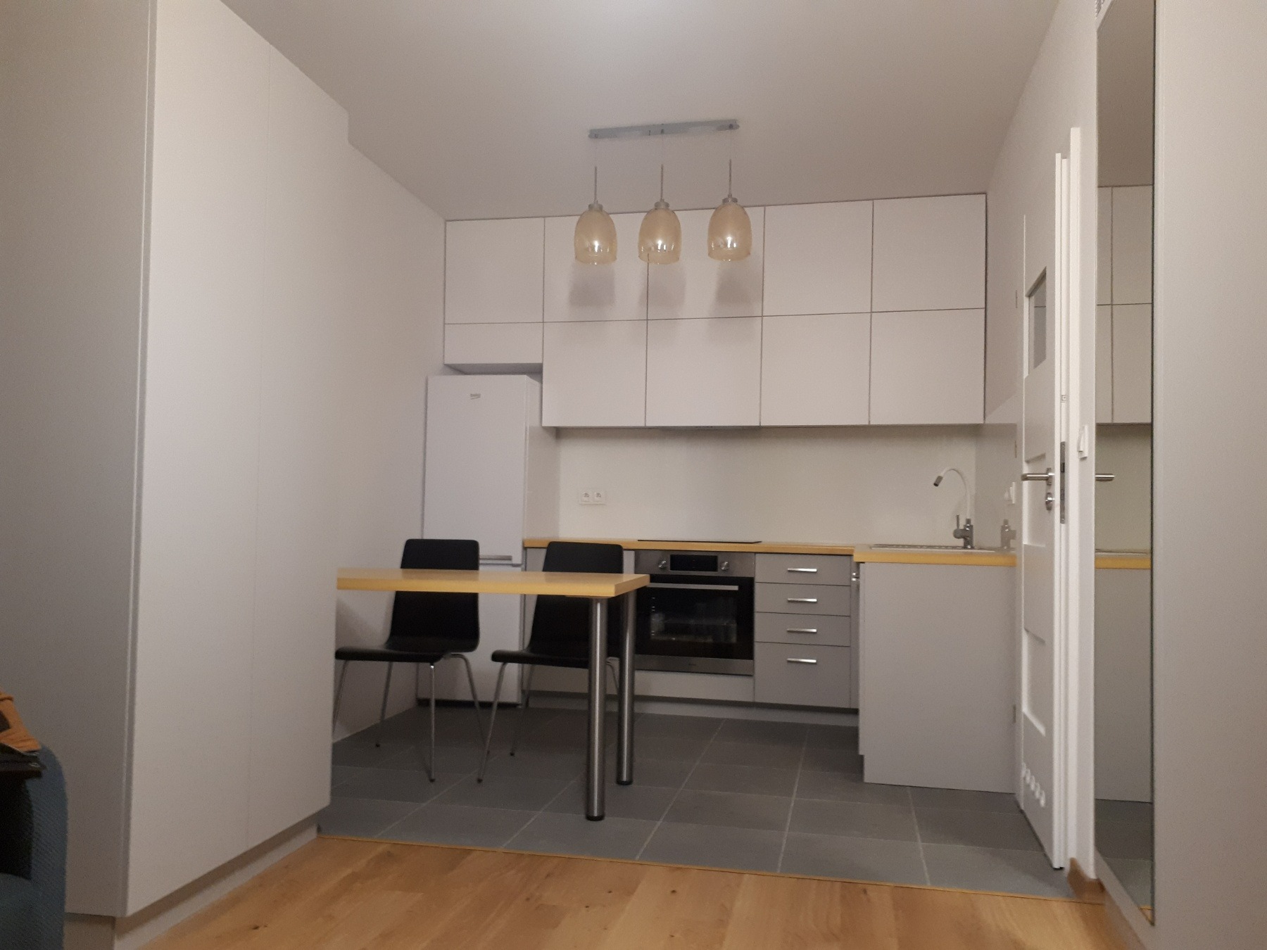 Jasna kuchnia w minimalistycznym stylu z białymi szafkami, szarymi blatami i podłogą, oświetlona trzema wiszącymi lampami z bursztynowego szkła, z widocznym stołem z dwoma krzesłami i częściowo...