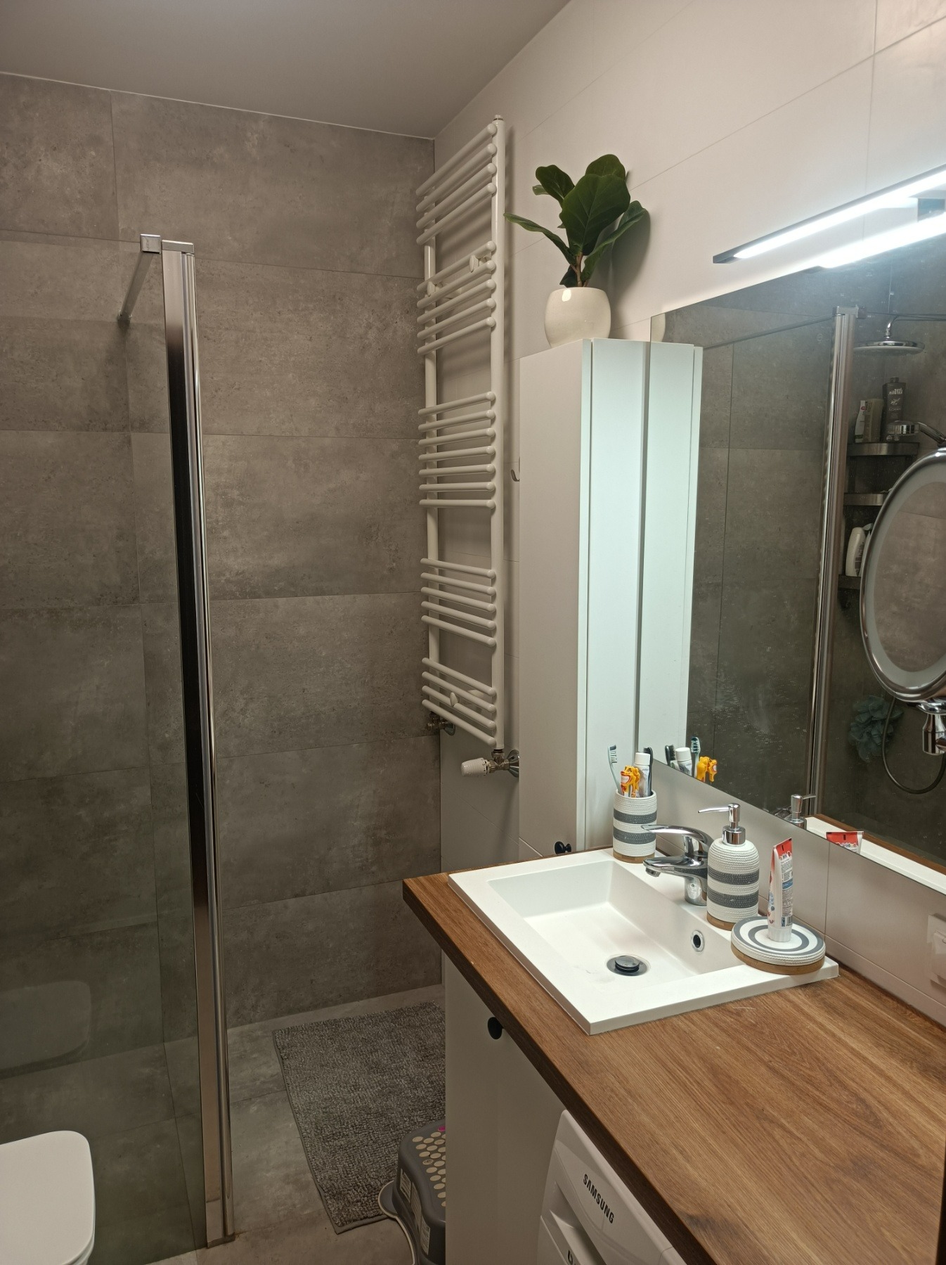 Nowoczesna łazienka z szarymi płytkami, kabiną prysznicową ze srebrnymi ramami, białym grzejnikiem drabinkowym, umywalką wpuszczaną w drewniany blat i wiszącą szafką z rośliną doniczkową na górze.