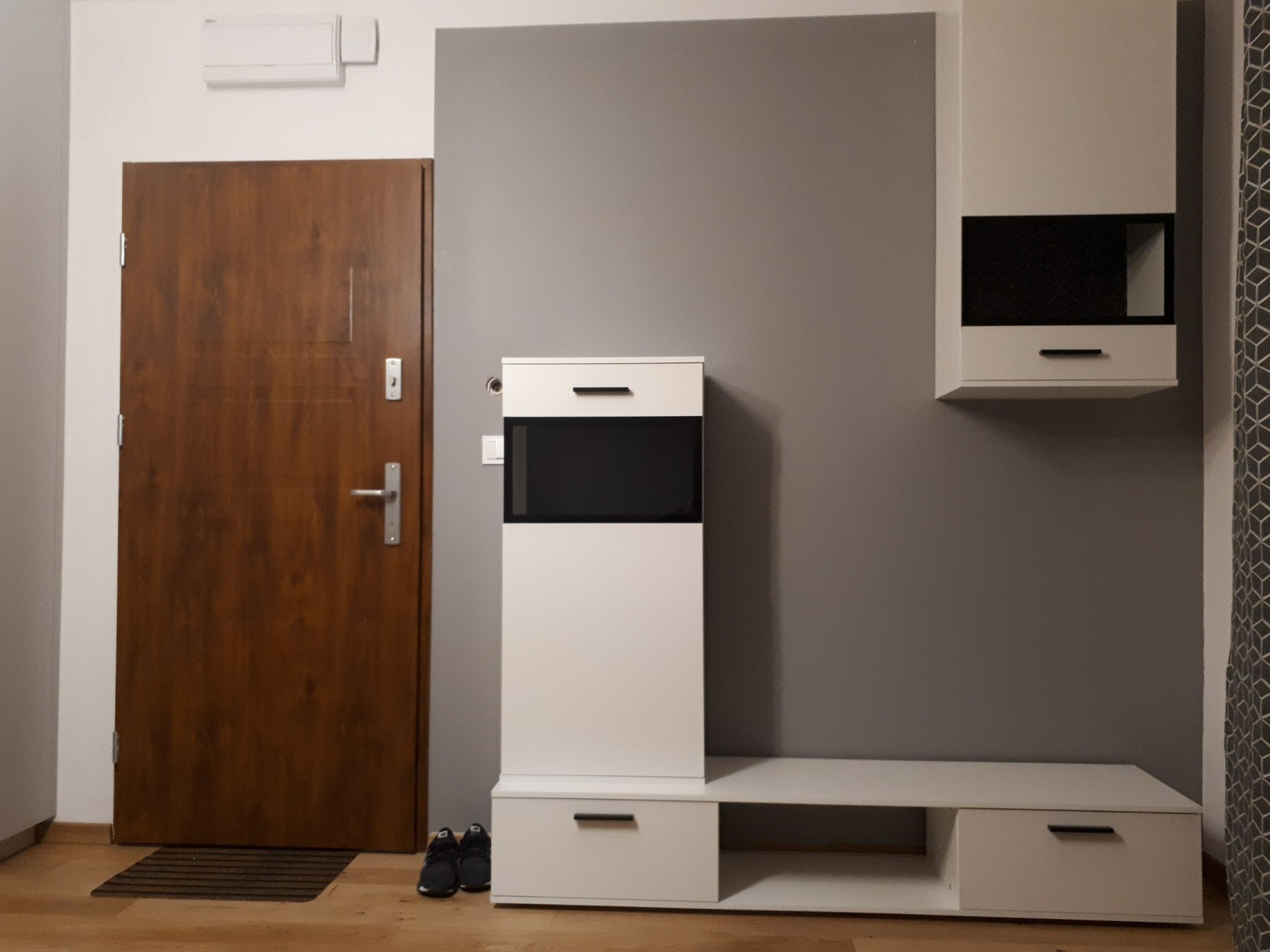 Nowoczesny przedpokój z brązowymi drzwiami, szarą ścianą i białymi meblami: szafka z czarnym frontem, wisząca szafka i niska komoda. Na podłodze leżą buty i wycieraczka.