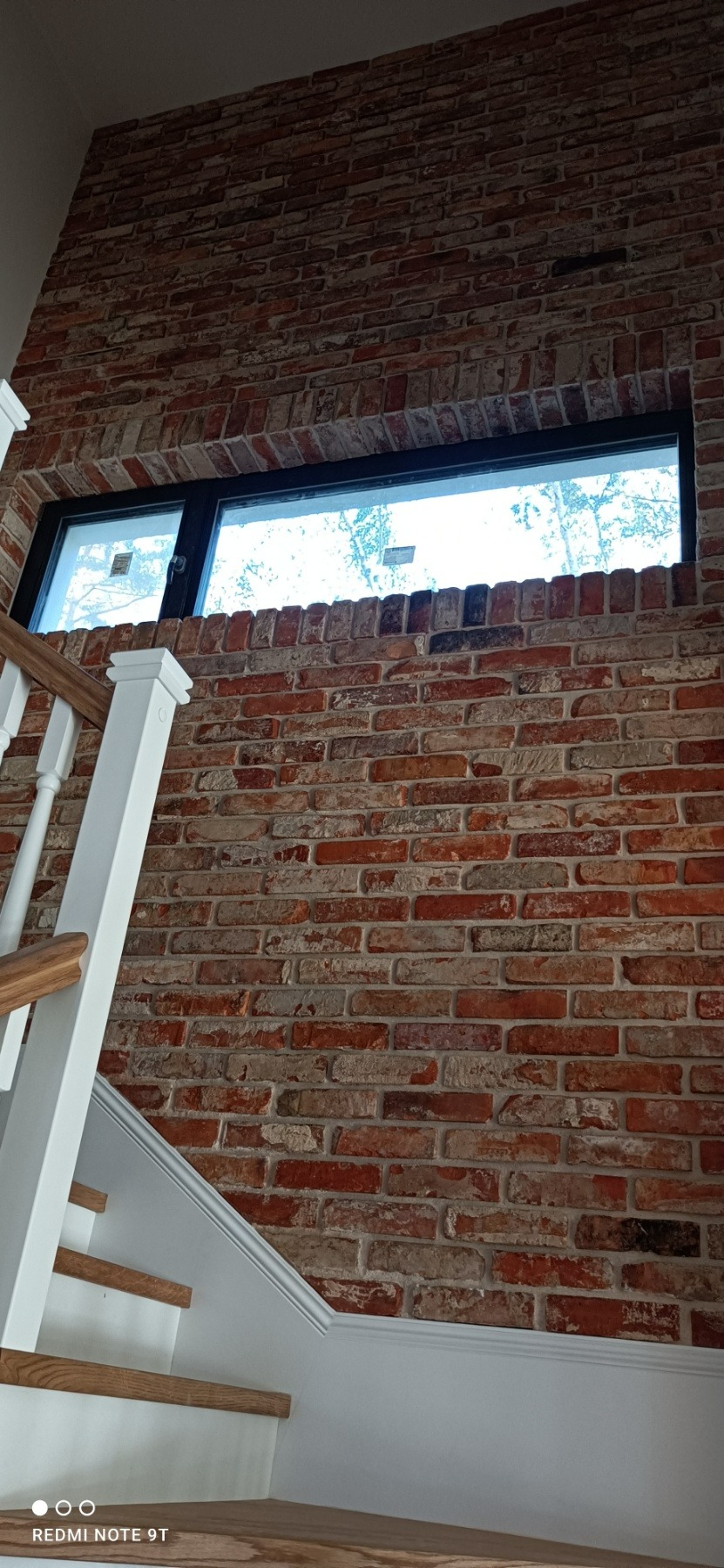 Ściana z cegły klinkierowej z oknem w czarnej ramie, widok na schody z białymi balustradami i drewnianymi stopniami.