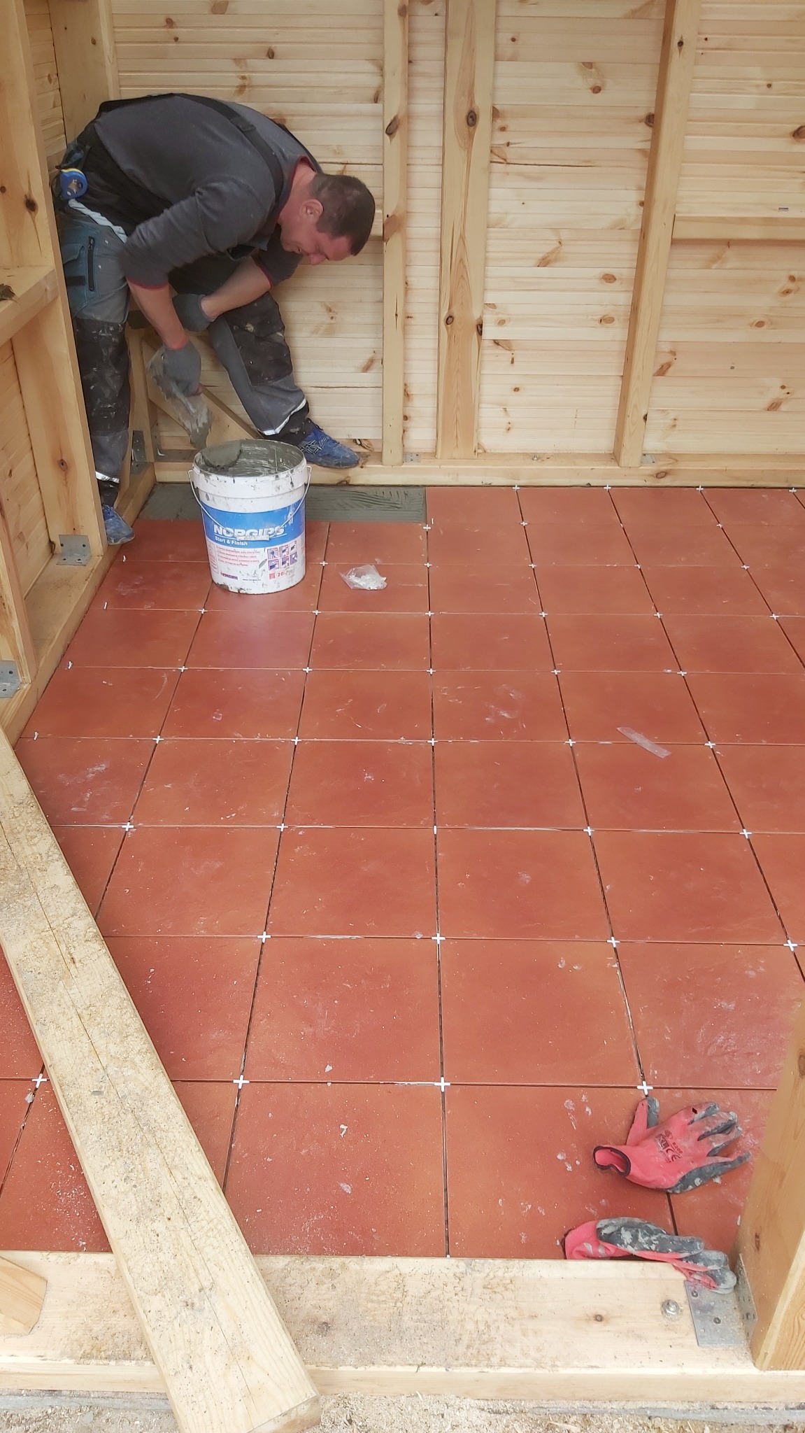 Glazurnik układa terakotę w drewnianej konstrukcji, widoczny kubeł z klejem Norgips i robocze rękawice na podłodze.