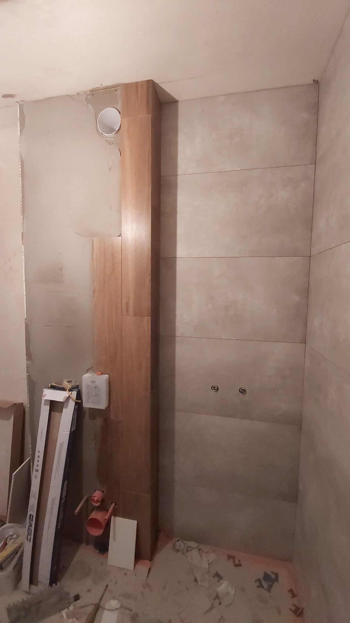 Fragment łazienki w trakcie remontu, widoczne ułożone płytki w odcieniach beżu, częściowo położone drewnopodobne płytki na pionowej zabudowie, wystające rury i niedokończone ściany, na podłodze...