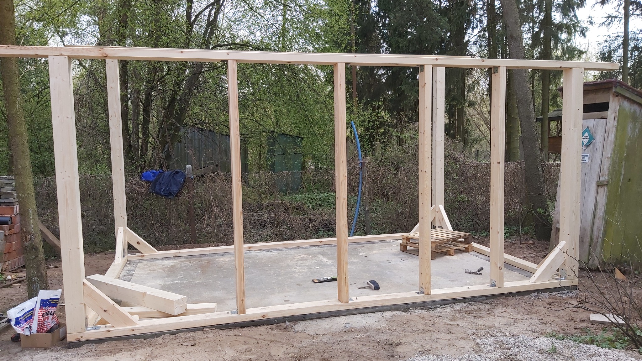 Drewniana konstrukcja szkieletowa budynku w trakcie budowy, widoczne słupy i belki, na betonowej wylewce leżą narzędzia. W tle zieleń drzew.