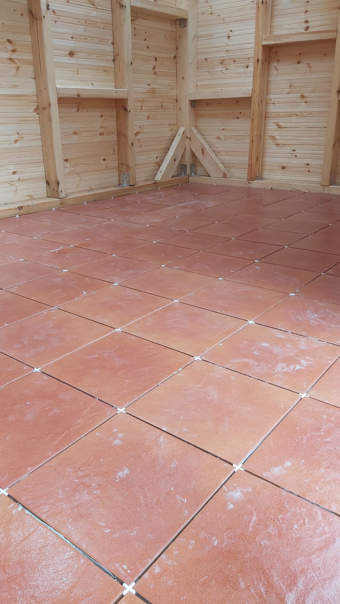 Podłoga z terakoty w trakcie układania, widoczne krzyżyki dystansowe, drewniana konstrukcja ścian i sufitu w surowym stanie.