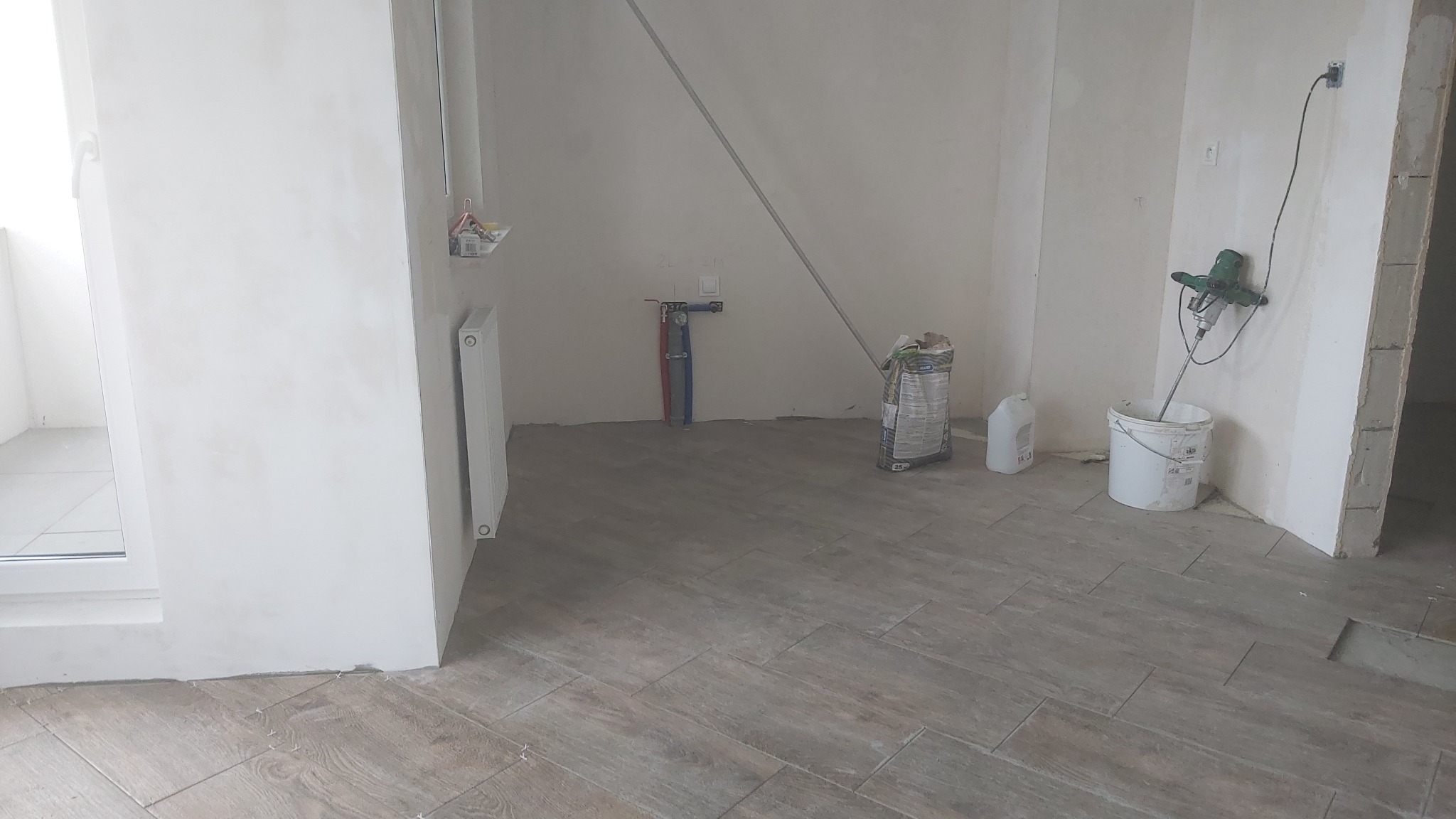 Wnętrze w trakcie prac wykończeniowych: położona podłoga z płytek imitujących drewno, ściany przygotowane pod gładzie, widoczne worki z zaprawą, mieszadło elektryczne w wiadrze, instalacja wodna...