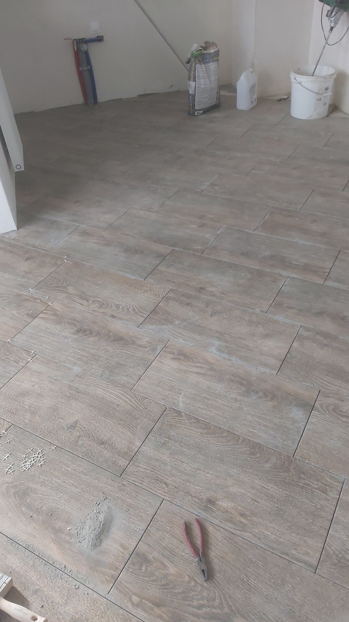 Ułożona podłoga z płytek imitujących drewno, widoczne krzyżyki dystansowe, paczka kleju, wiadro i szczypce na podłodze w trakcie prac wykończeniowych.