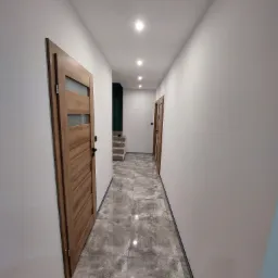 Wykończony korytarz w domu z drewnianymi drzwiami z szybami, szarą podłogą z płytek imitujących kamień i białymi ścianami, oświetlony punktowymi lampami sufitowymi.