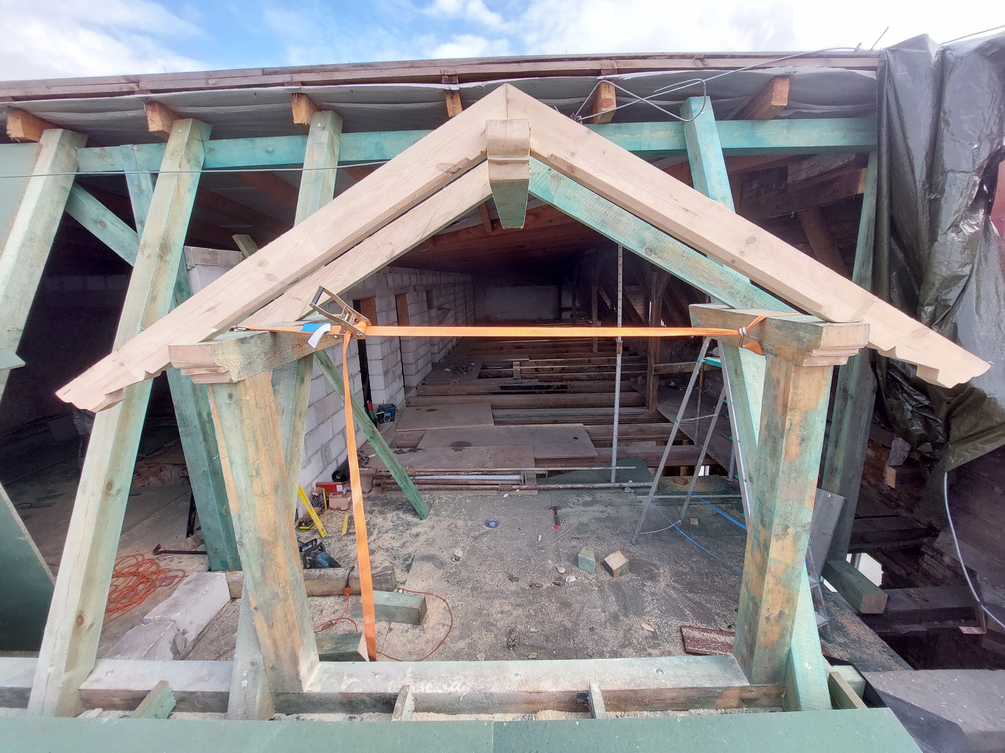 Konstrukcja drewniana dachu w trakcie budowy, widoczne narzędzia i materiały budowlane, niebieskie niebo w tle.