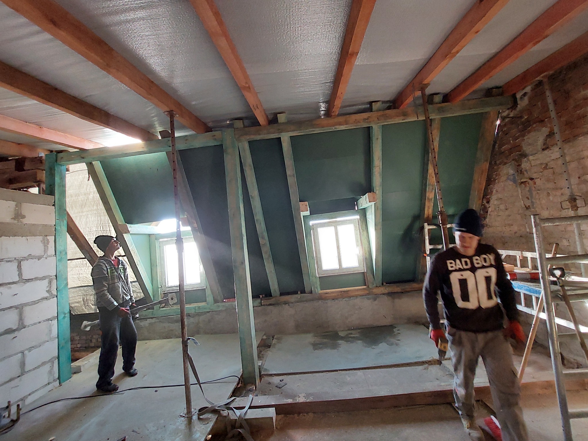 Adaptacja poddasza: Dwóch mężczyzn w roboczych ubraniach pracuje nad konstrukcją drewnianą pod ocieplenie, widoczne okna dachowe i folia izolacyjna na suficie.
