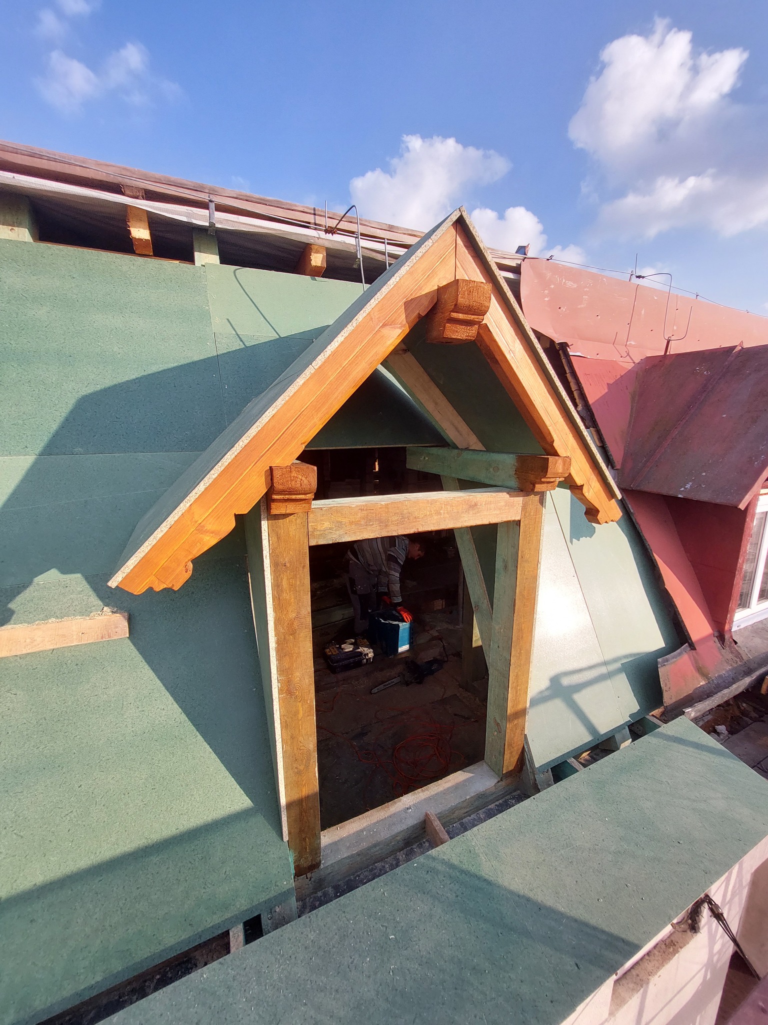 Montaż drewnianej lukarny w dachu pokrytym zieloną płytą OSB, widoczne narzędzia i częściowo sylwetka pracownika w środku