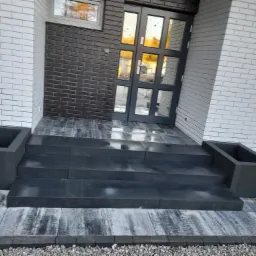 Nowoczesne schody wejściowe z ciemnej kostki brukowej prowadzące do przeszklonych drzwi wejściowych, z donicami po bokach, na tle białej i grafitowej elewacji z cegły klinkierowej.