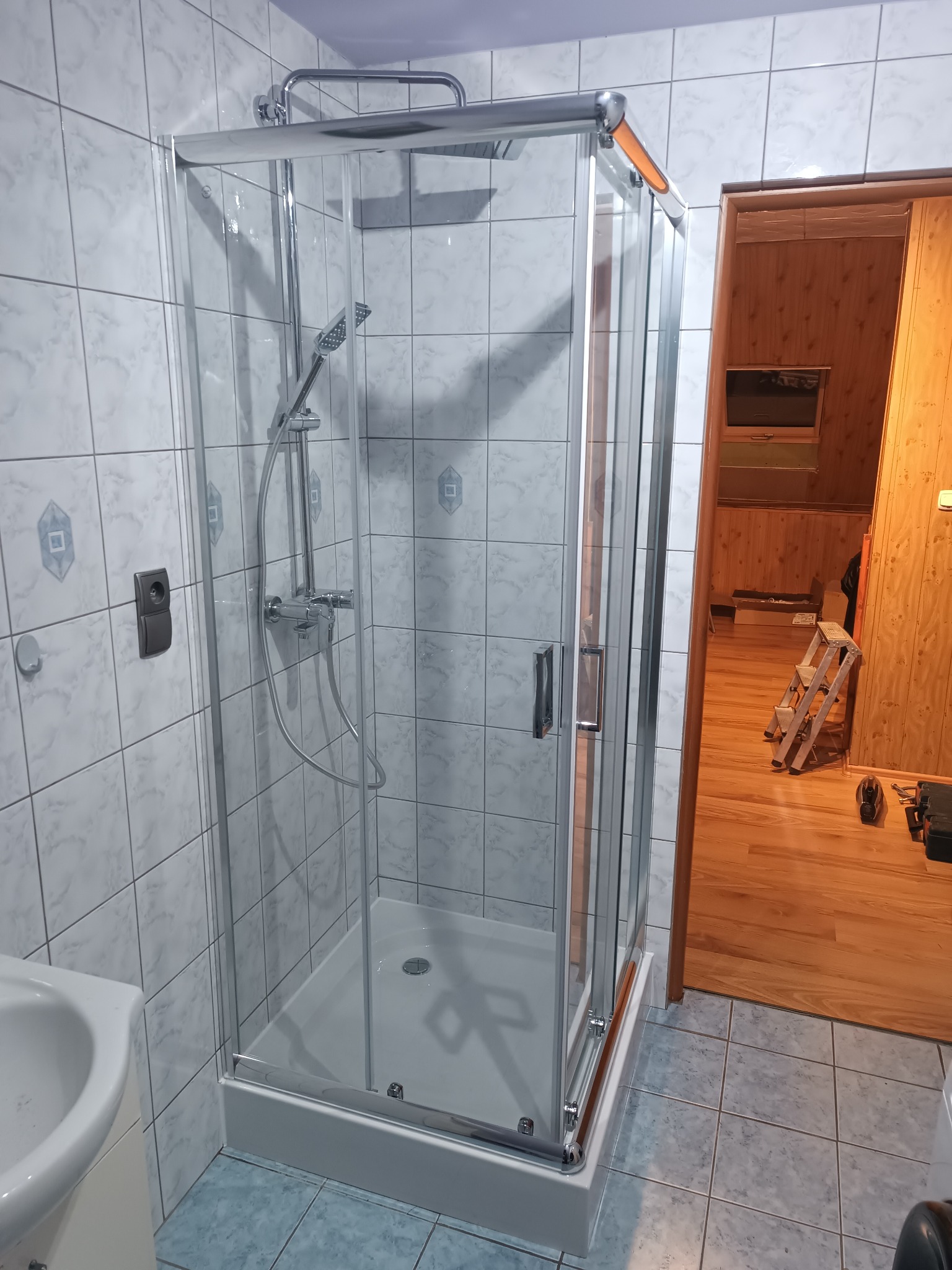 Szklana kabina prysznicowa z chromowanymi elementami w łazience wyłożonej białymi płytkami, widoczny fragment umywalki i otwarte drzwi do pomieszczenia z drewnianą drabiną.