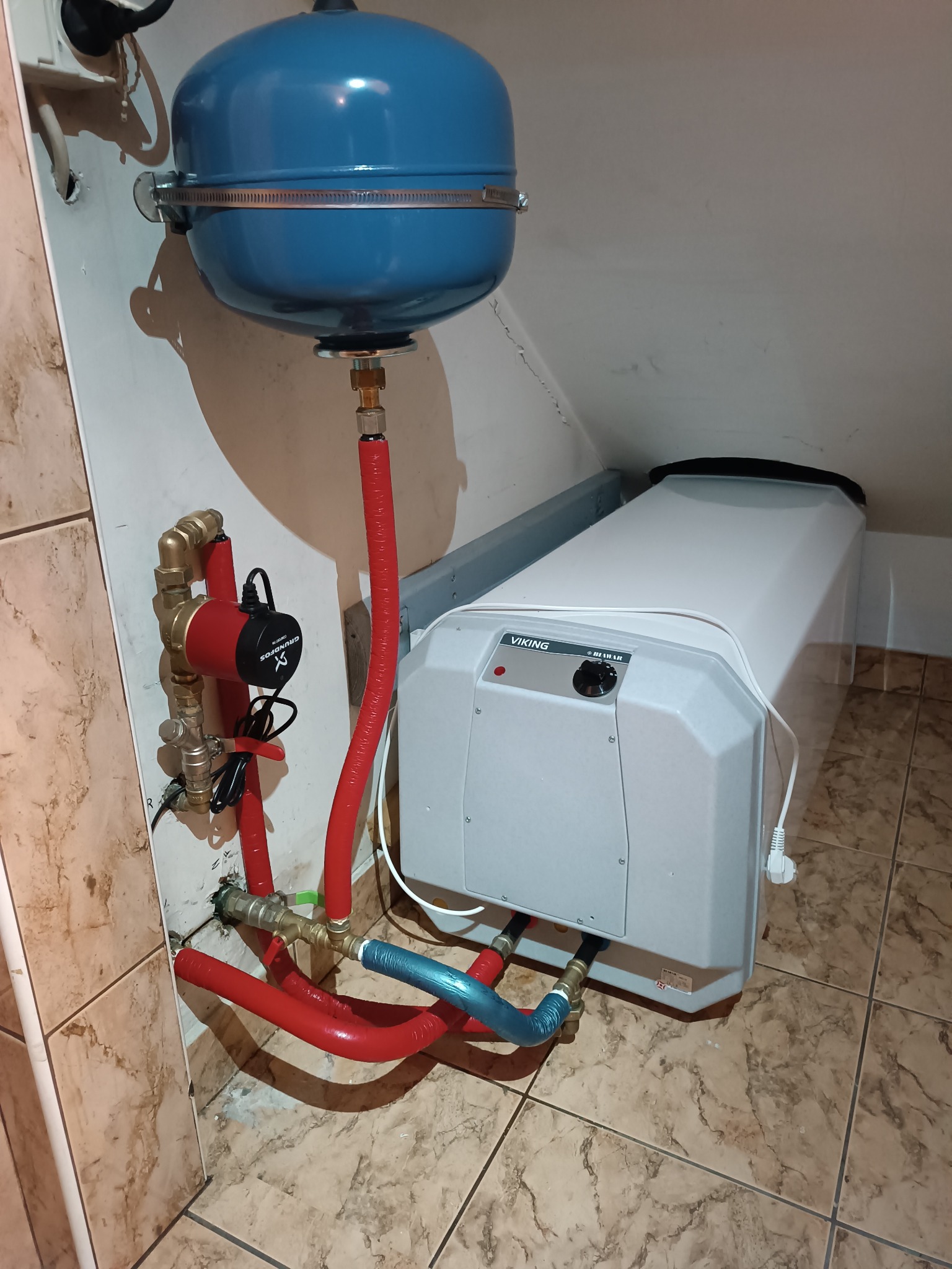 Kompaktowy piec grzewczy Viking z osprzętem: niebieski zbiornik wyrównawczy, czerwona pompa obiegowa Grundfos, rury w izolacji czerwonej i niebieskiej, zamontowany w pomieszczeniu ze skośnym...