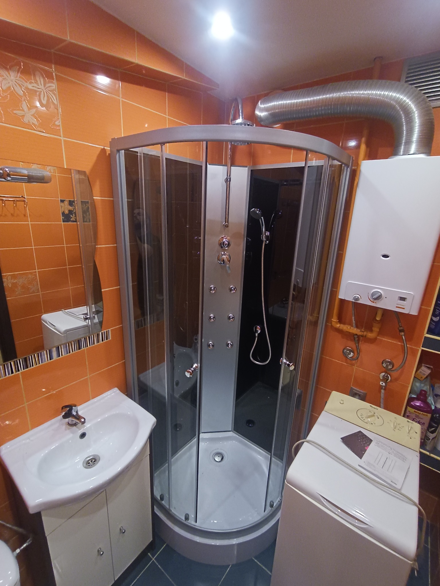 Narożna kabina prysznicowa z hydromasażem w łazience z pomarańczowymi płytkami, widoczny bojler gazowy z podłączeniem i umywalka z szafką.