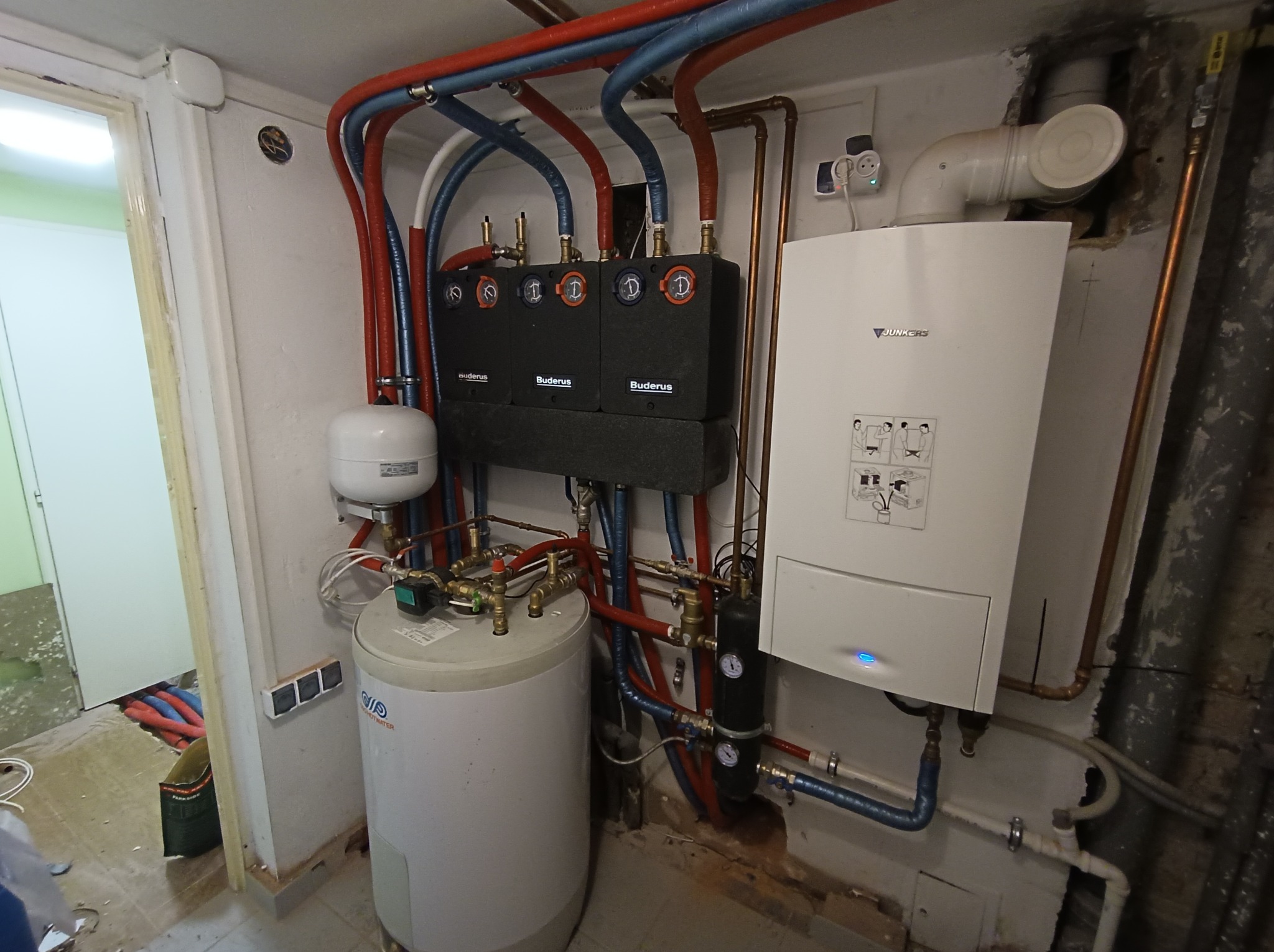 Kotłownia z kotłem Junkers, zasobnikiem ciepłej wody i rozdzielaczami Buderus, widoczne liczne rury miedziane i plastikowe w kolorach czerwonym i niebieskim.