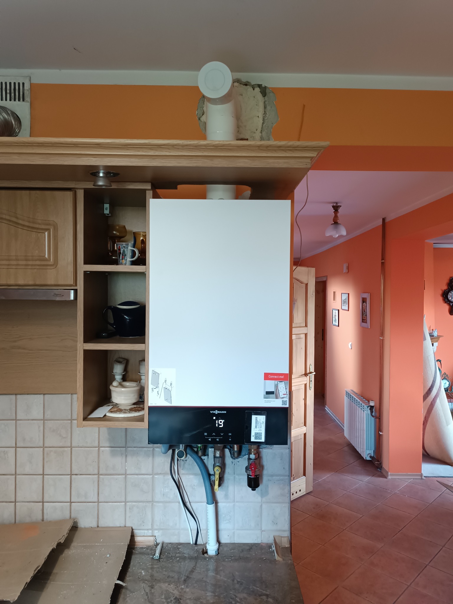 Nowy, biały piec gazowy Viessmann zamontowany w kuchni, z widocznymi podłączeniami hydraulicznymi i gazowymi. W tle pomarańczowe ściany i fragment grzejnika.