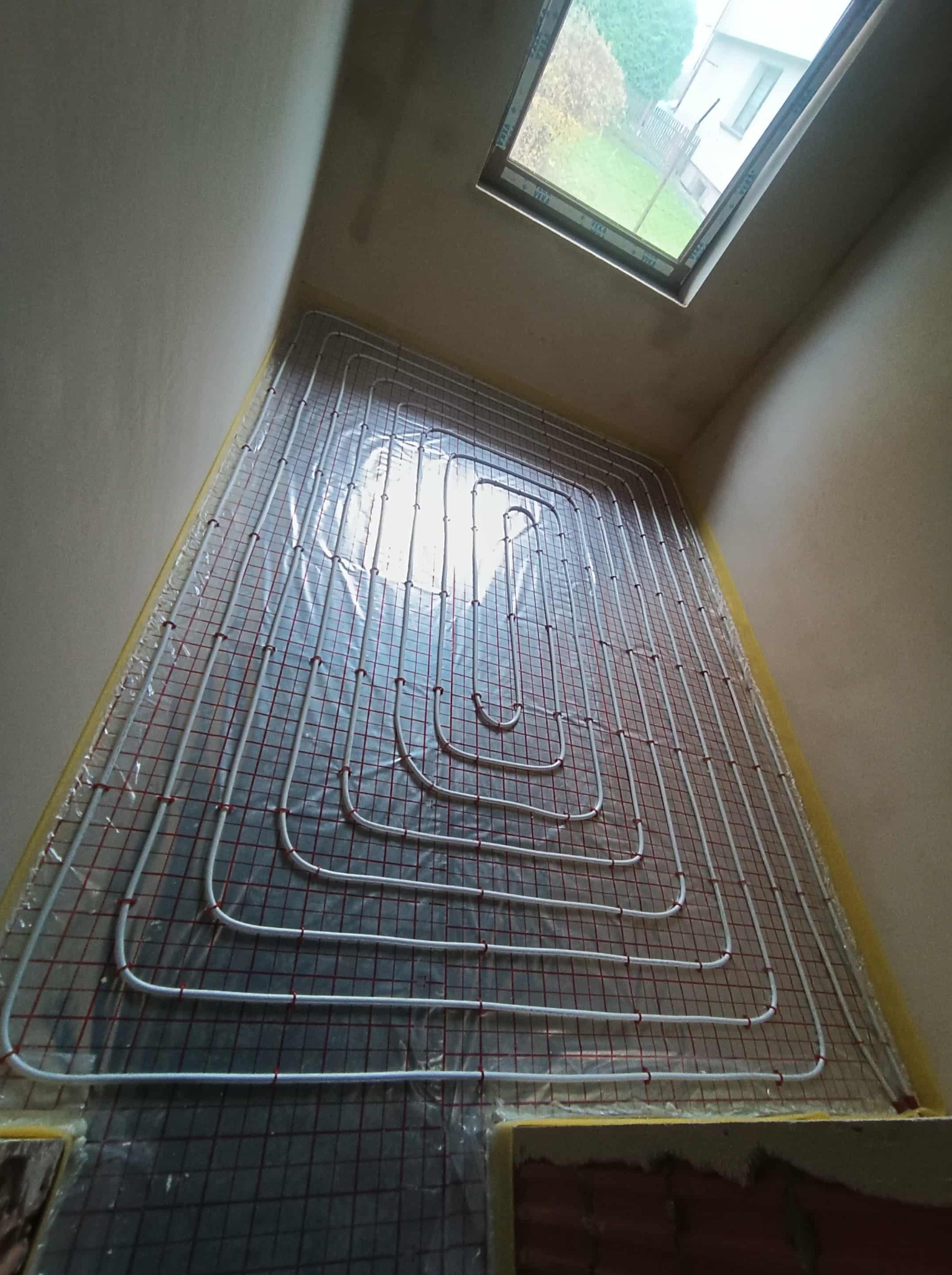 Białe rury ogrzewania podłogowego ułożone na czarnej macie, widok z góry, z oknem dachowym w tle. Instalacja zabezpieczona folią przy ścianach.