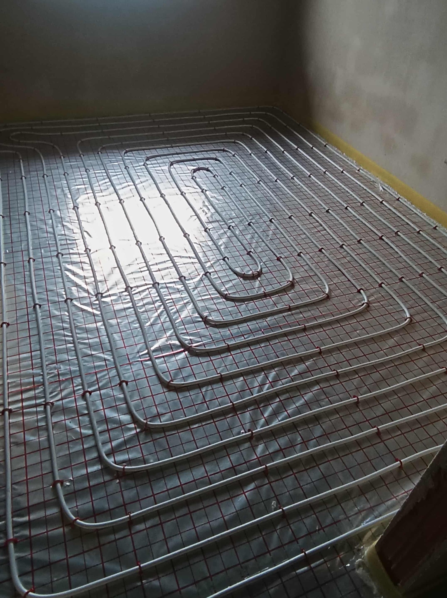 Białe rury ogrzewania podłogowego ułożone na folii z czerwoną siatką w surowym wnętrzu. Widok z góry, instalacja przed wylaniem posadzki.