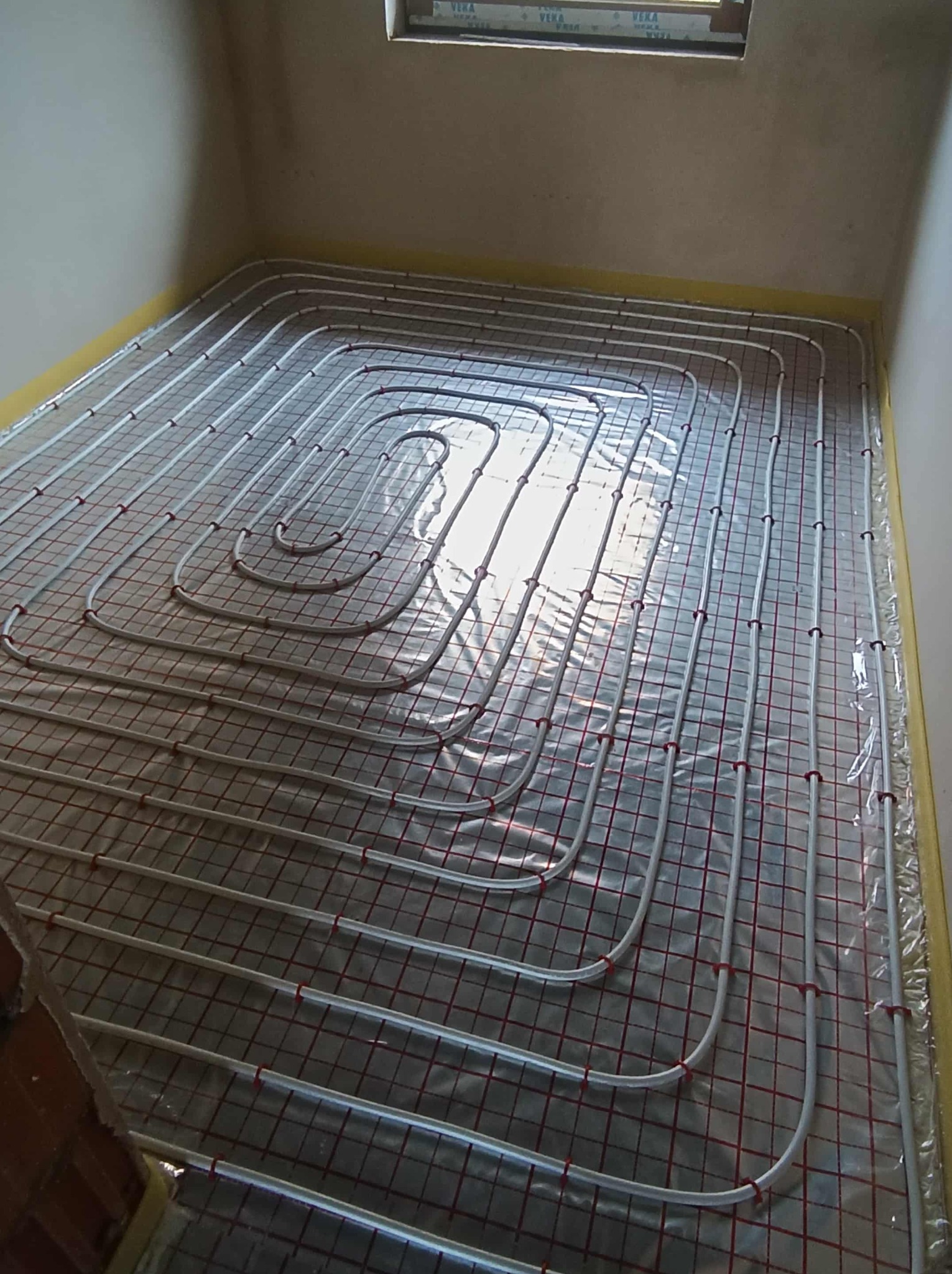 Białe rury ogrzewania podłogowego ułożone na folii z czerwoną siatką, widoczne okno w tle. Instalacja przed wylaniem posadzki.
