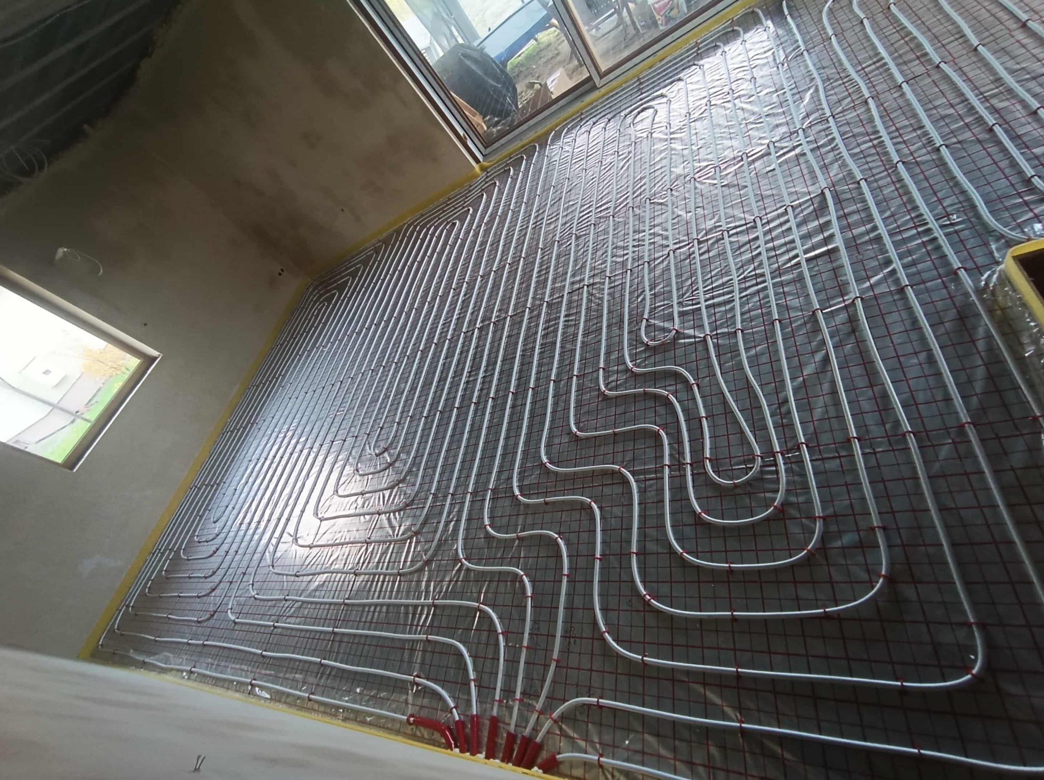 Białe rury ogrzewania podłogowego ułożone na folii i siatce w surowym wnętrzu. Widoczne okna i fragmenty ścian w trakcie prac wykończeniowych.