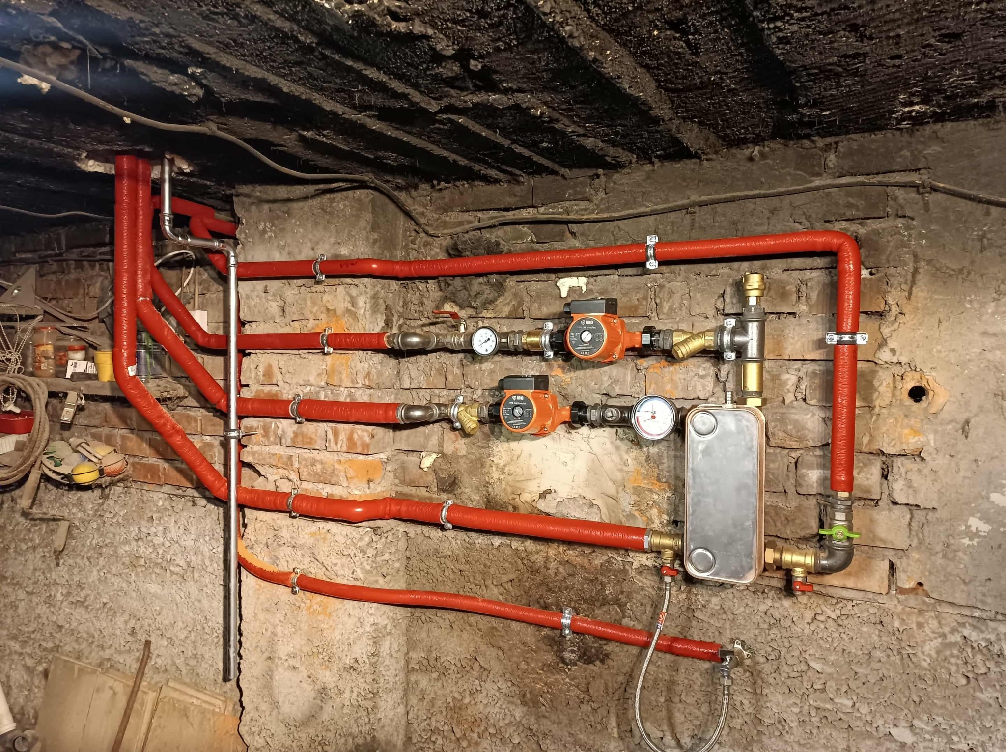 Czerwone rury grzewcze z izolacją, pompy obiegowe i wymiennik ciepła zamontowane na starej, ceglanej ścianie w piwnicy. Instalacja po modernizacji.