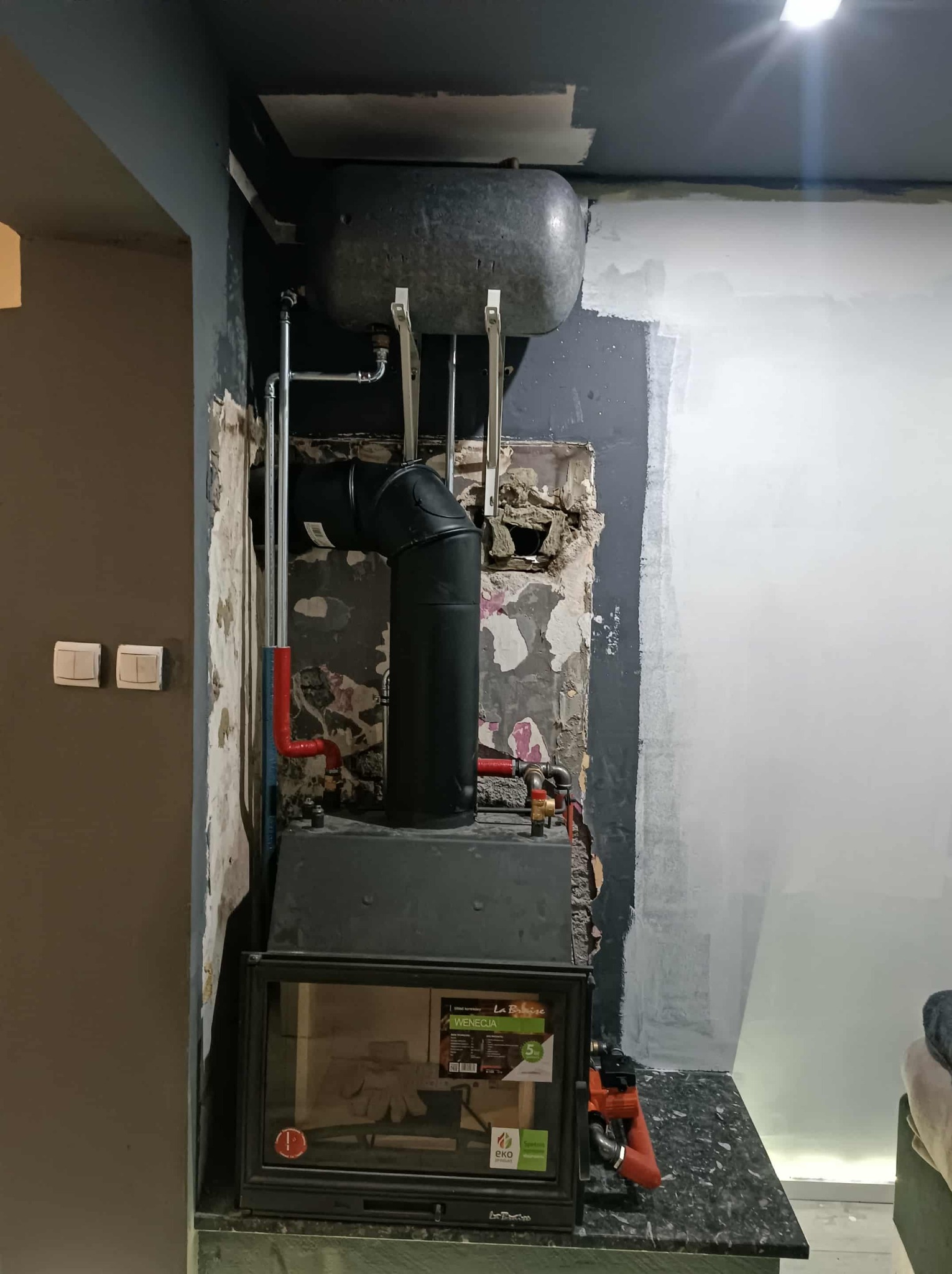 Czarny piec z instalacją grzewczą w trakcie montażu, widoczne rury i zbiornik. Ściany w trakcie wykończenia, surowy stan. Ujęcie z bliska, wnętrze pomieszczenia.