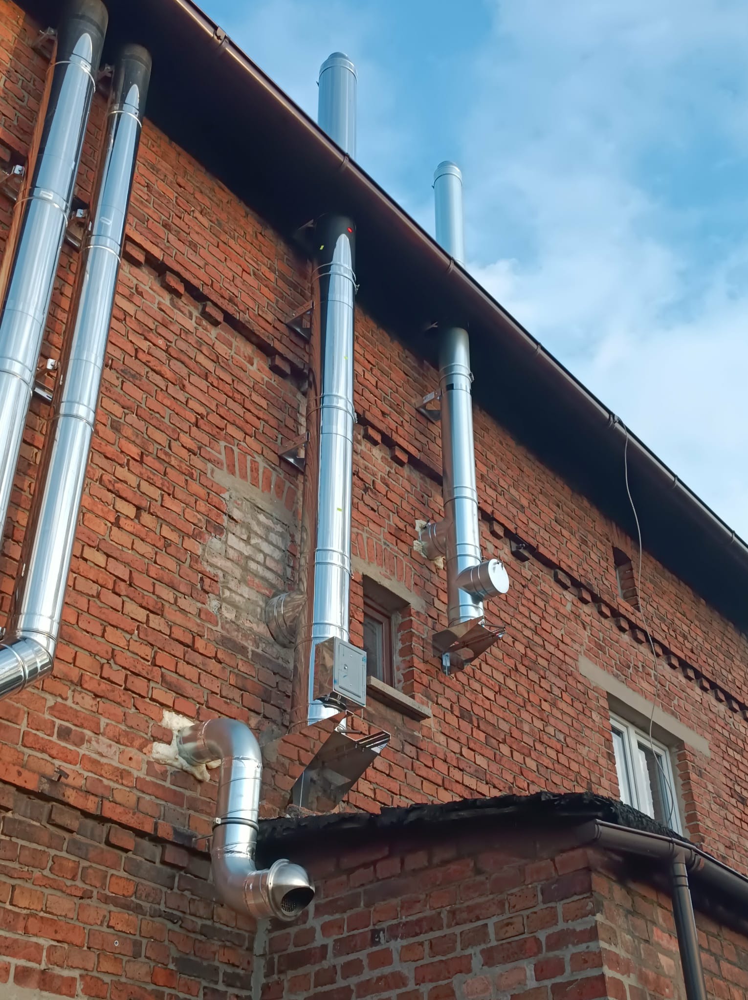 Zewnętrzna instalacja kominowa z połyskujących, metalowych rur na tle ceglanej ściany budynku i fragmentu błękitnego nieba. Widok z dołu, podkreślający pionowy układ.