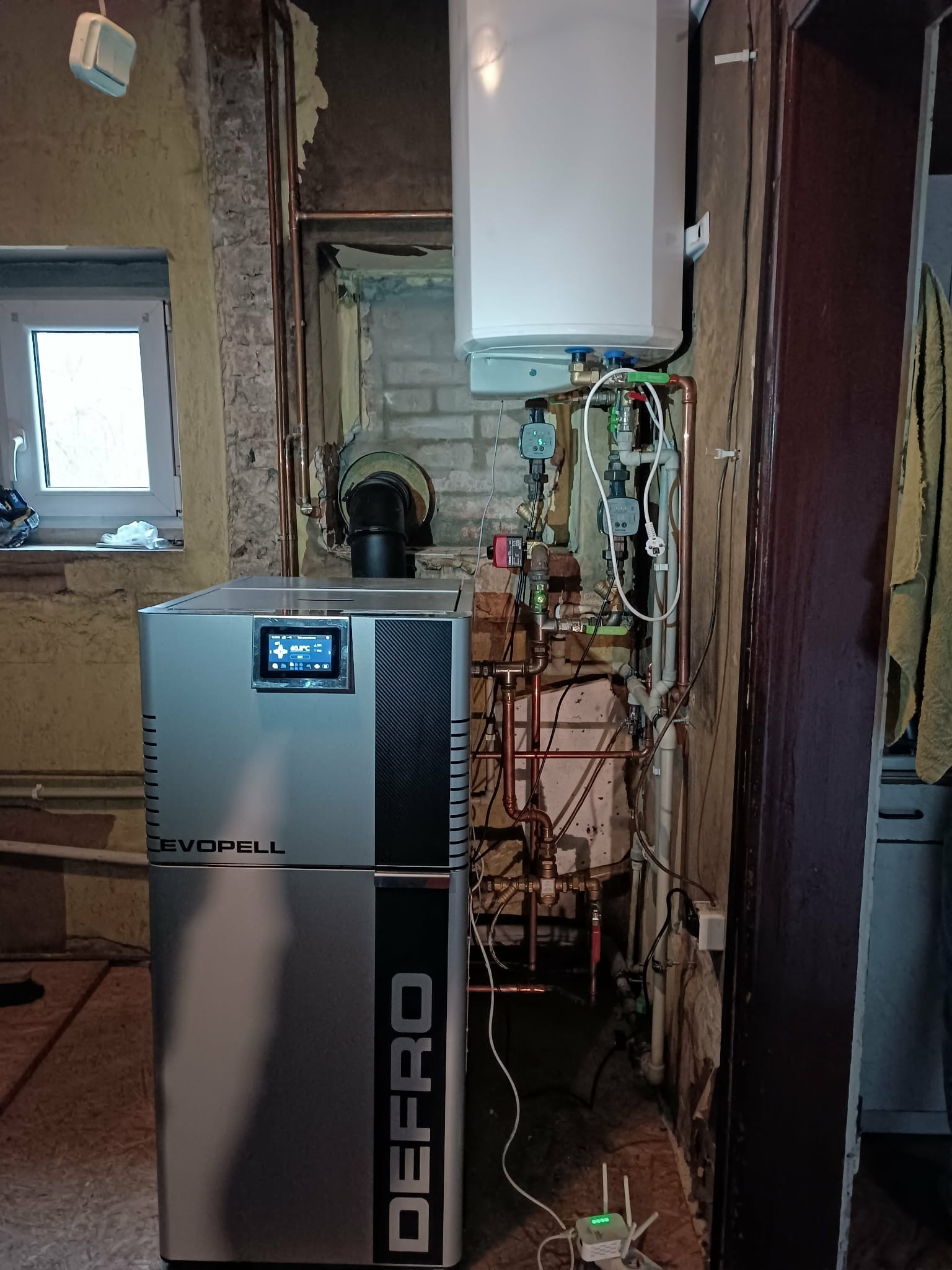 Nowoczesny piec Defro Evopell z ekranem dotykowym, obok instalacja grzewcza z miedzianymi rurami i białym zasobnikiem. Widoczne okno i fragment drzwi.
