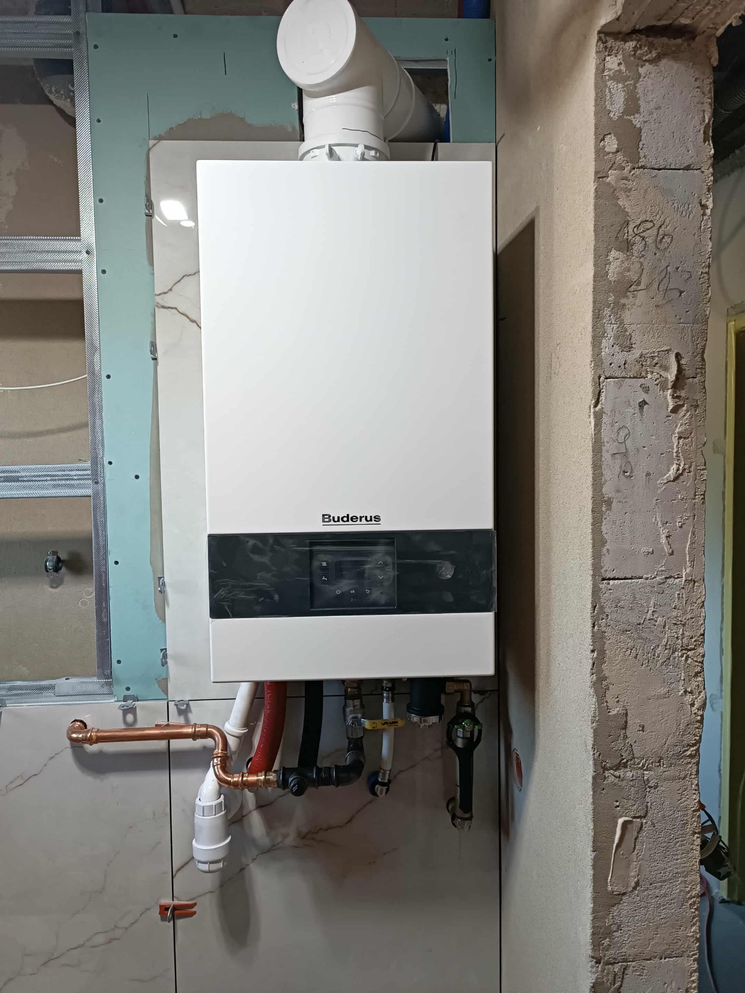 Biały piec gazowy Buderus z podłączeniami hydraulicznymi na tle surowej ściany i fragmentu konstrukcji. Widoczne miedziane rury i elementy instalacji.