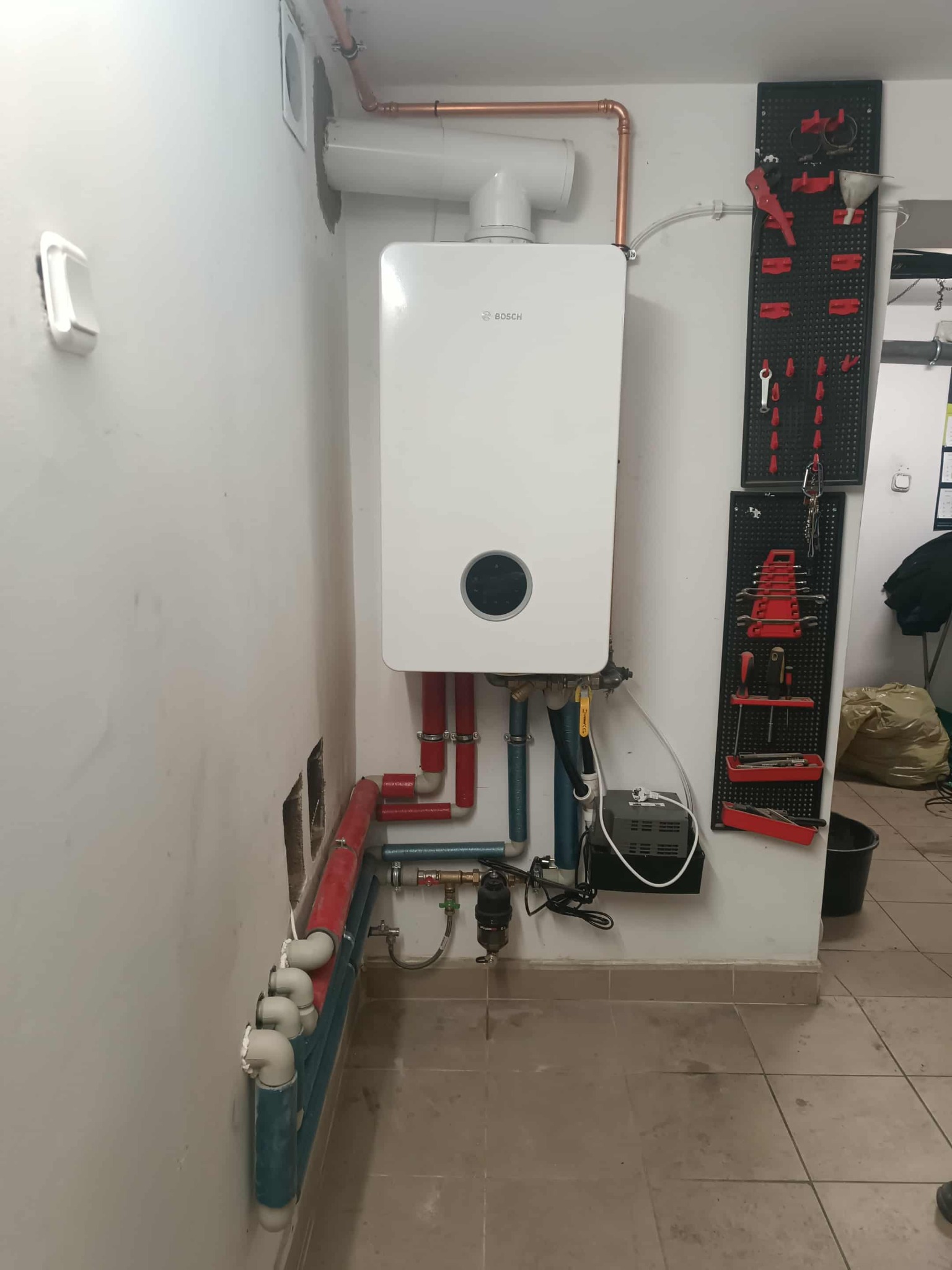 Biały piec gazowy Bosch zamontowany na ścianie z widocznymi rurami miedzianymi i PCV. Obok wieszak z narzędziami. Widoczne ślady po kuciu w ścianie.
