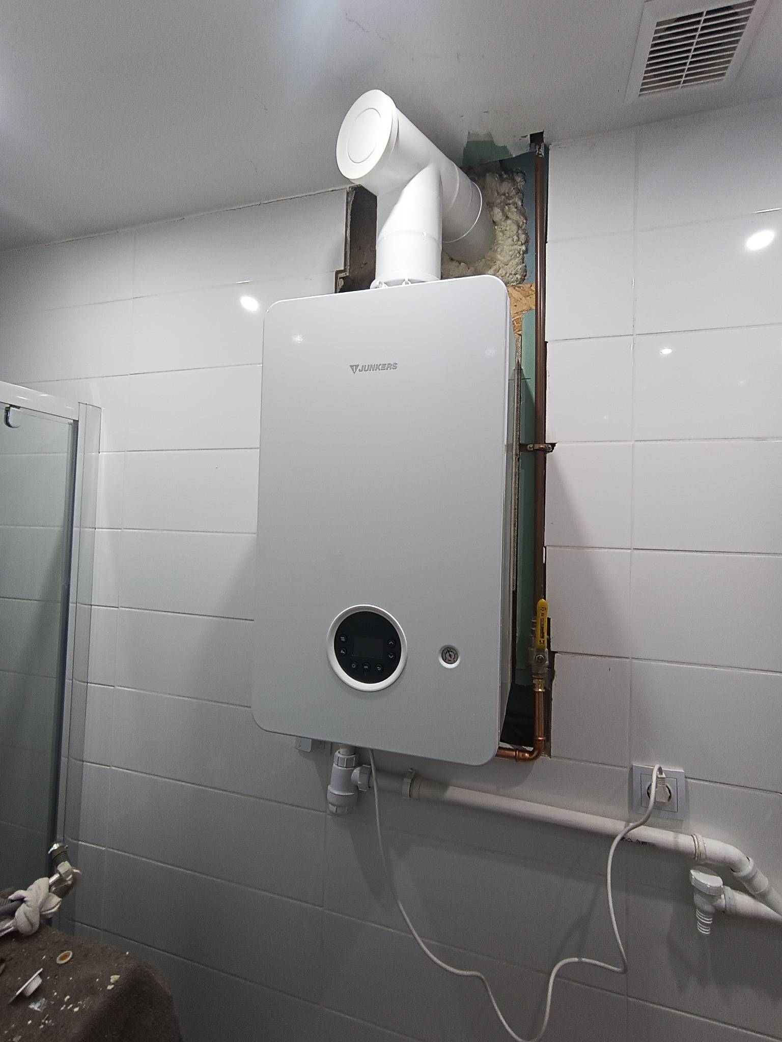 Biały piec gazowy Junkers zamontowany na kafelkowanej ścianie w łazience, widoczne rury i instalacja, fragment kabiny prysznicowej po lewej stronie.