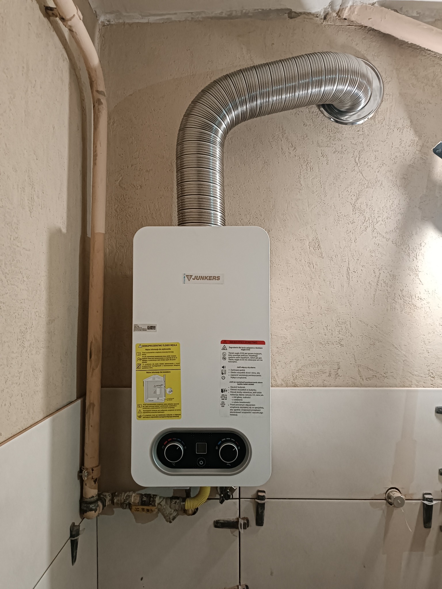 Biały, wiszący piec gazowy Junkers z podłączoną, elastyczną rurą odprowadzającą spaliny, zamontowany na kafelkowanej ścianie w pomieszczeniu. Widoczne rury i elementy instalacji.