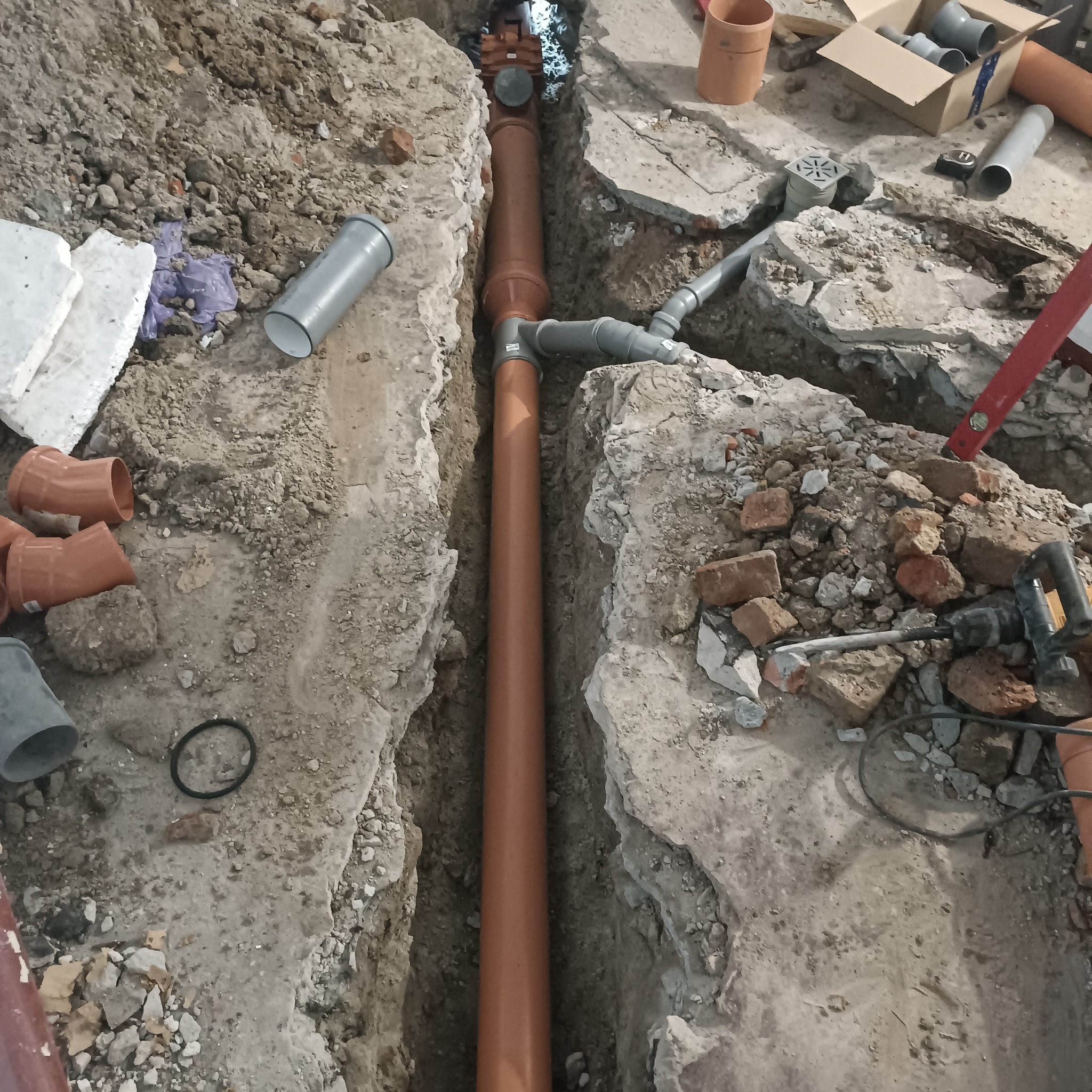 Wykop z ułożoną brązową rurą kanalizacyjną w ziemi. Widoczne narzędzia, gruz i elementy instalacji. Ujęcie z góry, ukazujące etap prac instalacyjnych.