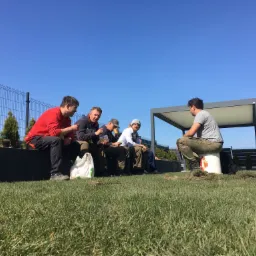 Grupa mężczyzn w odzieży roboczej odpoczywa i spożywa posiłek na skraju trawnika, w tle widoczny płot, tuje i pergola.