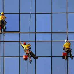 Trzech alpinist&oacute;w w kaskach, na linach, myje szklaną fasadę budynku biurowego w słoneczny dzień. Widoczne wiaderka i sprzęt do mycia okien.
