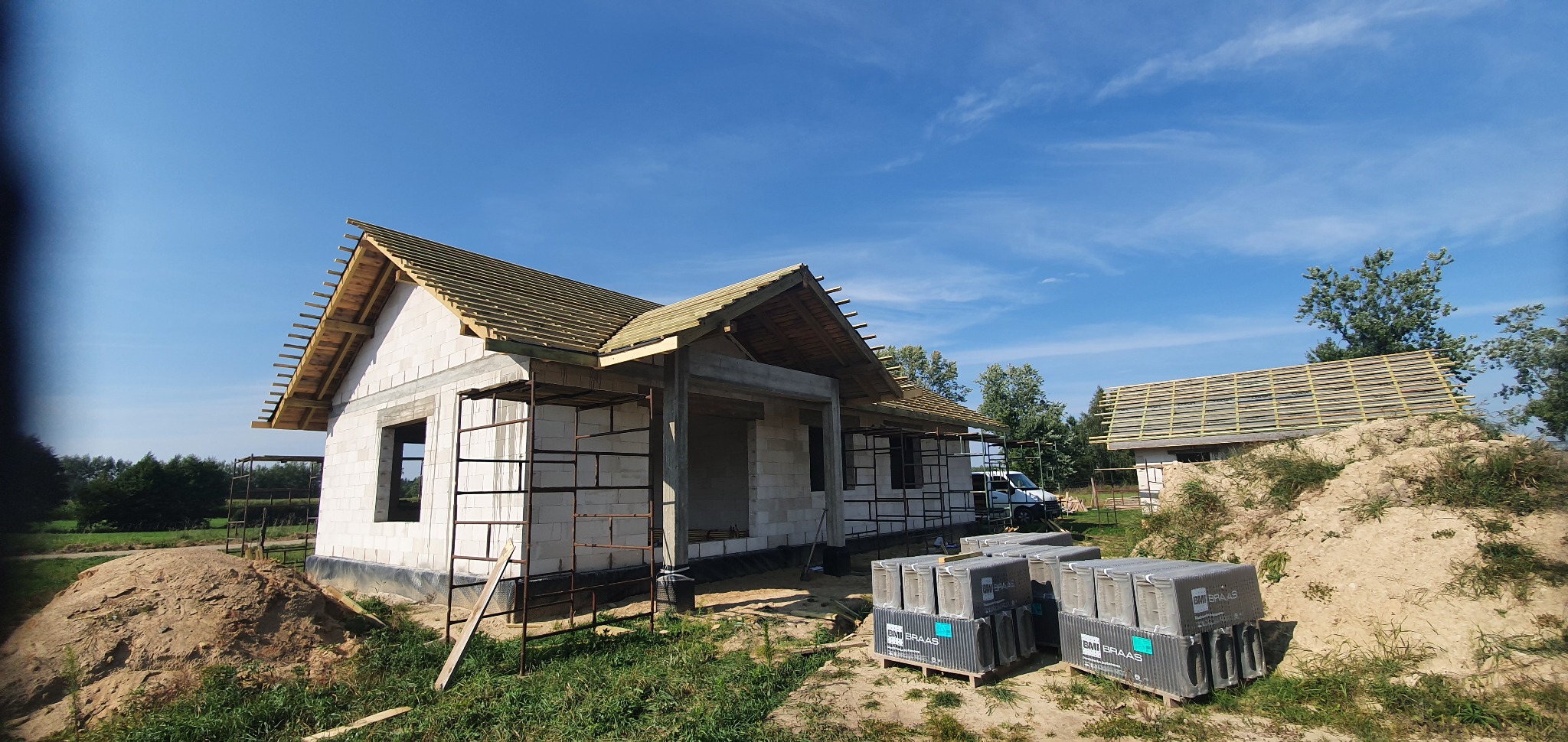 Budowa domu jednorodzinnego z widoczną konstrukcją dachu, na tle błękitnego nieba z delikatnymi chmurami, z paletami dachówek BMI Braas na pierwszym planie.