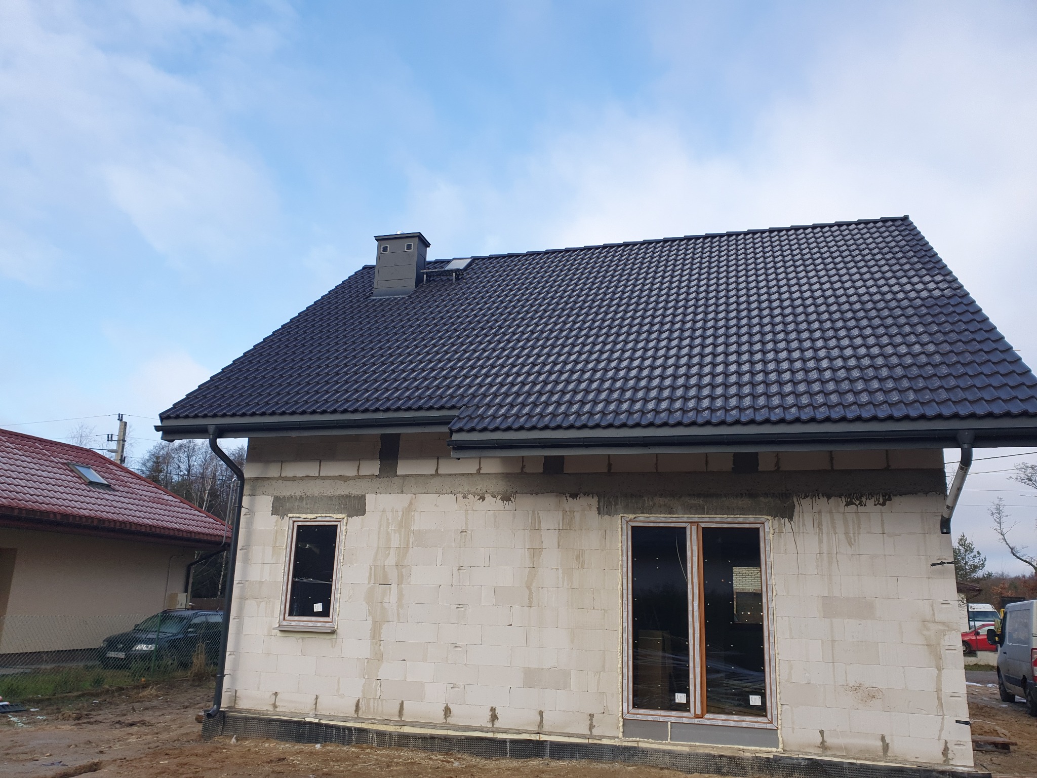 Nowy, grafitowy dach z blachodachówki modułowej na surowym budynku z betonu komórkowego, widoczne okna w stanie surowym, rynny i komin.
