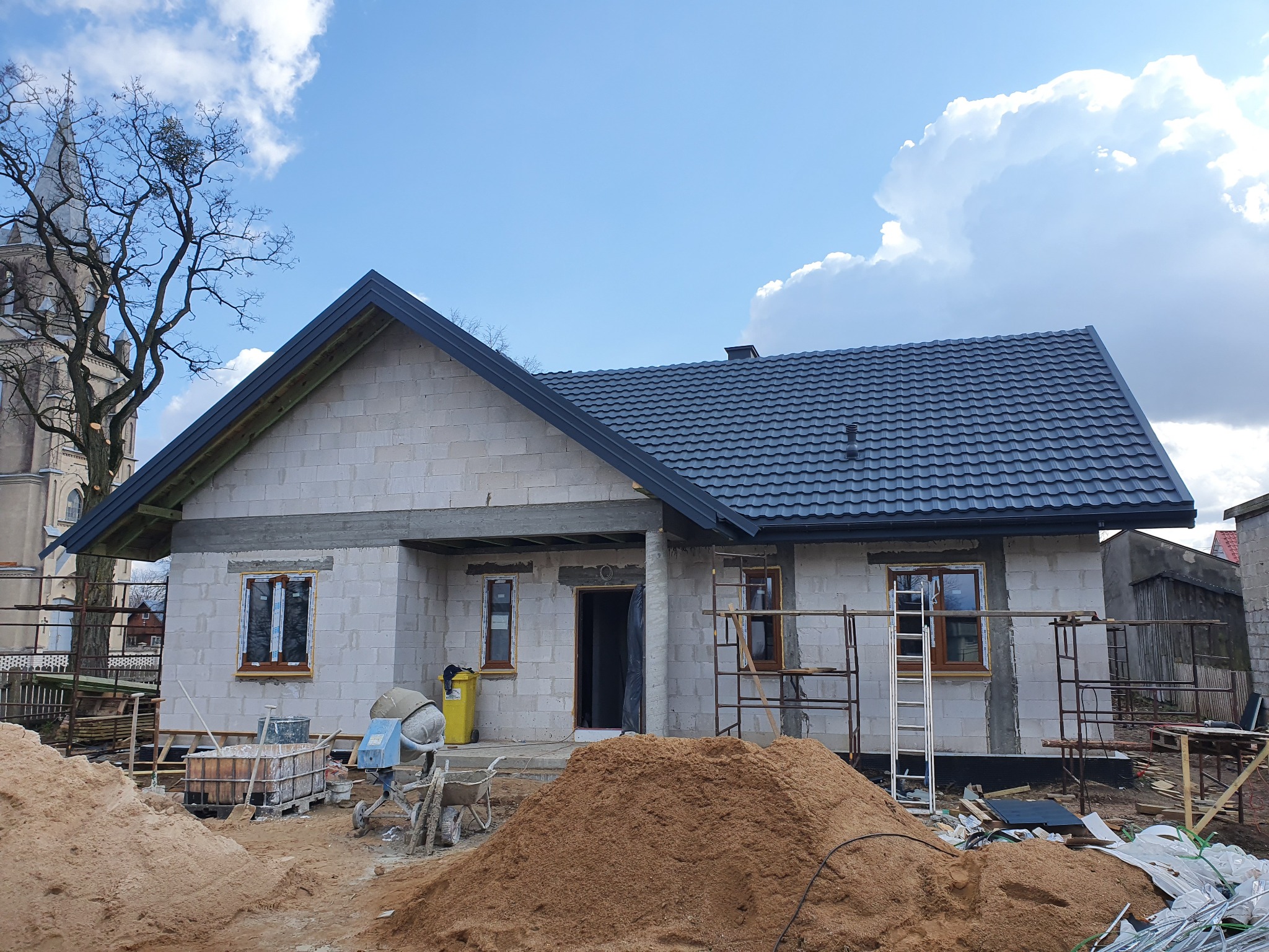 Surowy budynek jednorodzinny w trakcie budowy, z nowym, ciemnoszarym dachem z dachówki, widoczne rusztowania, piasek, betoniarka i kościół w tle.