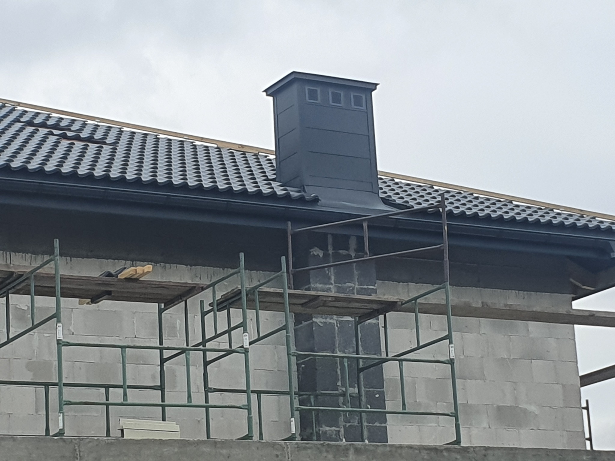 Nowy, grafitowy komin z obróbką blacharską na dachu z ciemnej dachówki, widoczny z rusztowania na tle szarej ściany budynku w trakcie budowy.