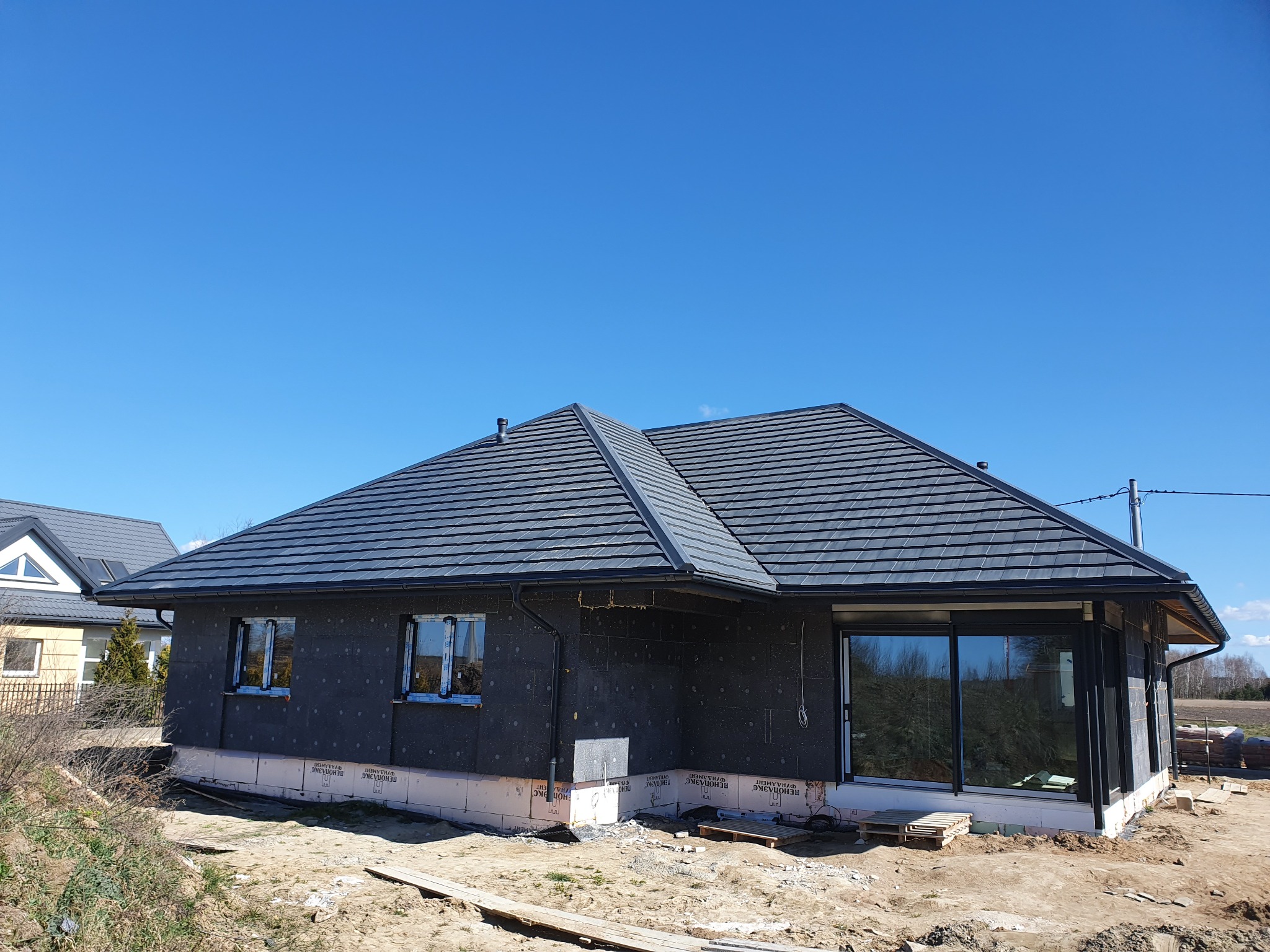 Dom jednorodzinny w trakcie budowy, z zamontowaną szarą dachówką ceramiczną i ociepleniem ścian zewnętrznych, widoczne okna i przygotowany teren.