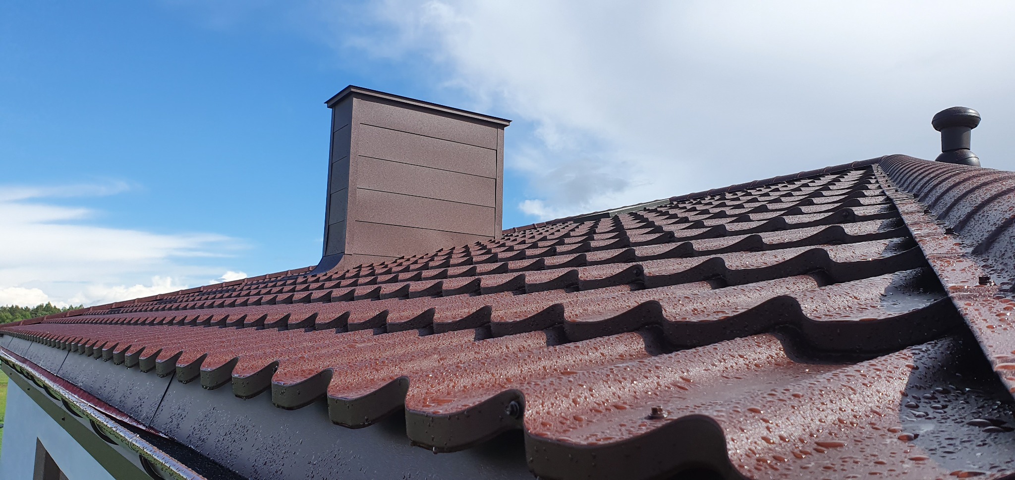 Czerwono-brązowe metalowe pokrycie dachowe z kroplami wody, widoczny komin z blachy oraz odpowietrznik, na tle błękitnego nieba z chmurami.